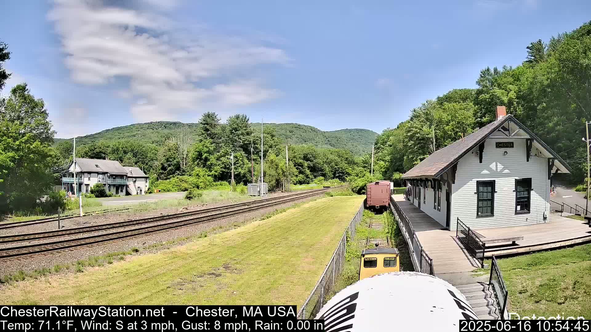 A white train station and a two-story building sit beside train tracks on a sunny day, with green hills in the background.