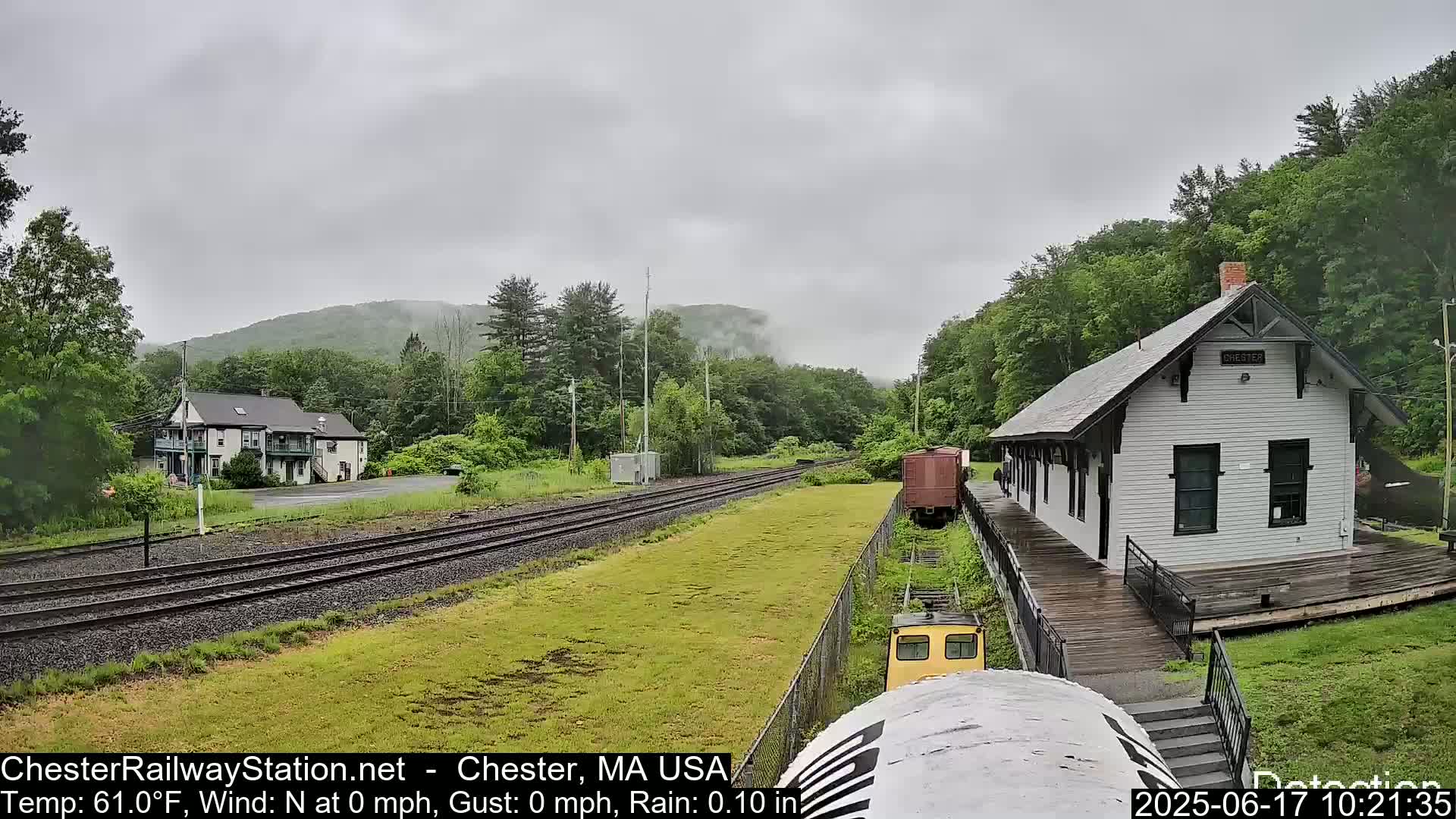 A rain-soaked train station sits beside railroad tracks, with a two-story building and lush green hills visible in the background under a cloudy sky.