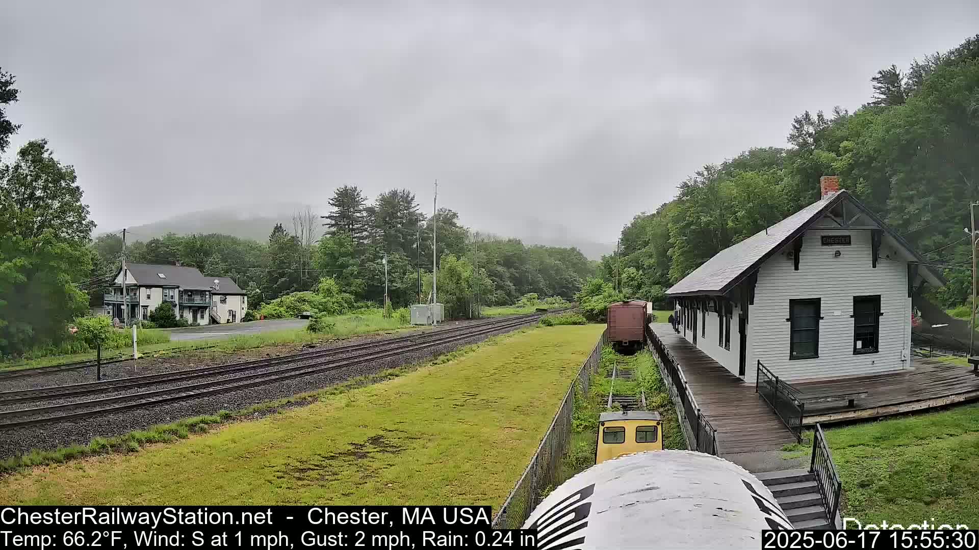 A small train approaches a white train station on a rainy day, with a two-story building visible in the background.