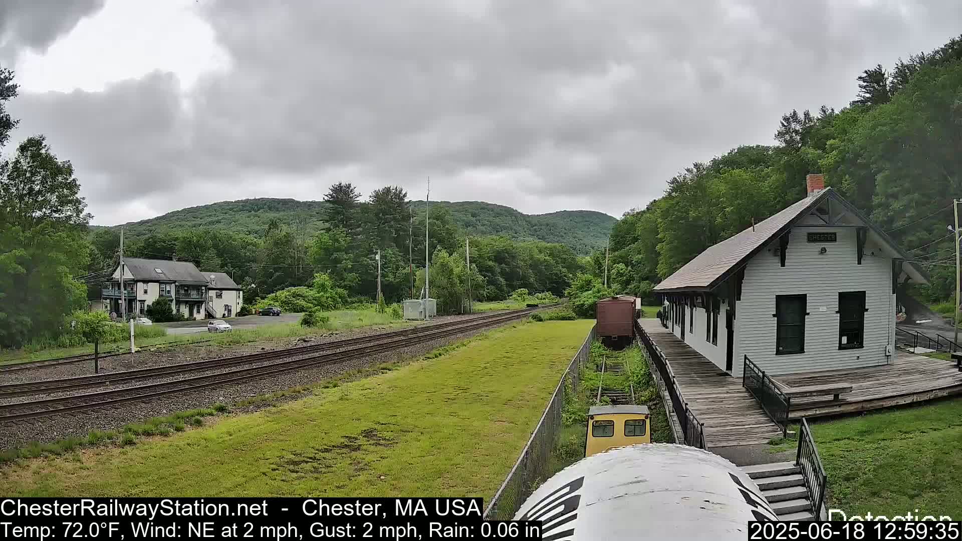 A white train station sits on a wooden platform beside train tracks, with a light rain falling under a cloudy sky, surrounded by green hills and trees.