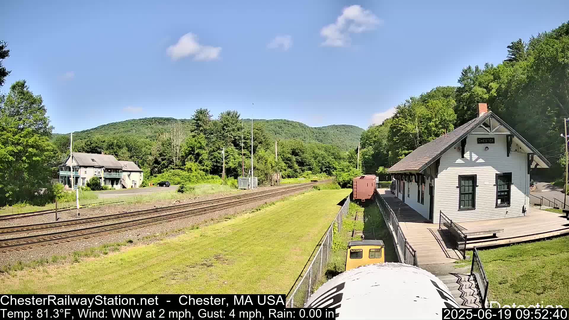 A train station and a two-story building sit beside train tracks on a sunny day, with green hills in the background.