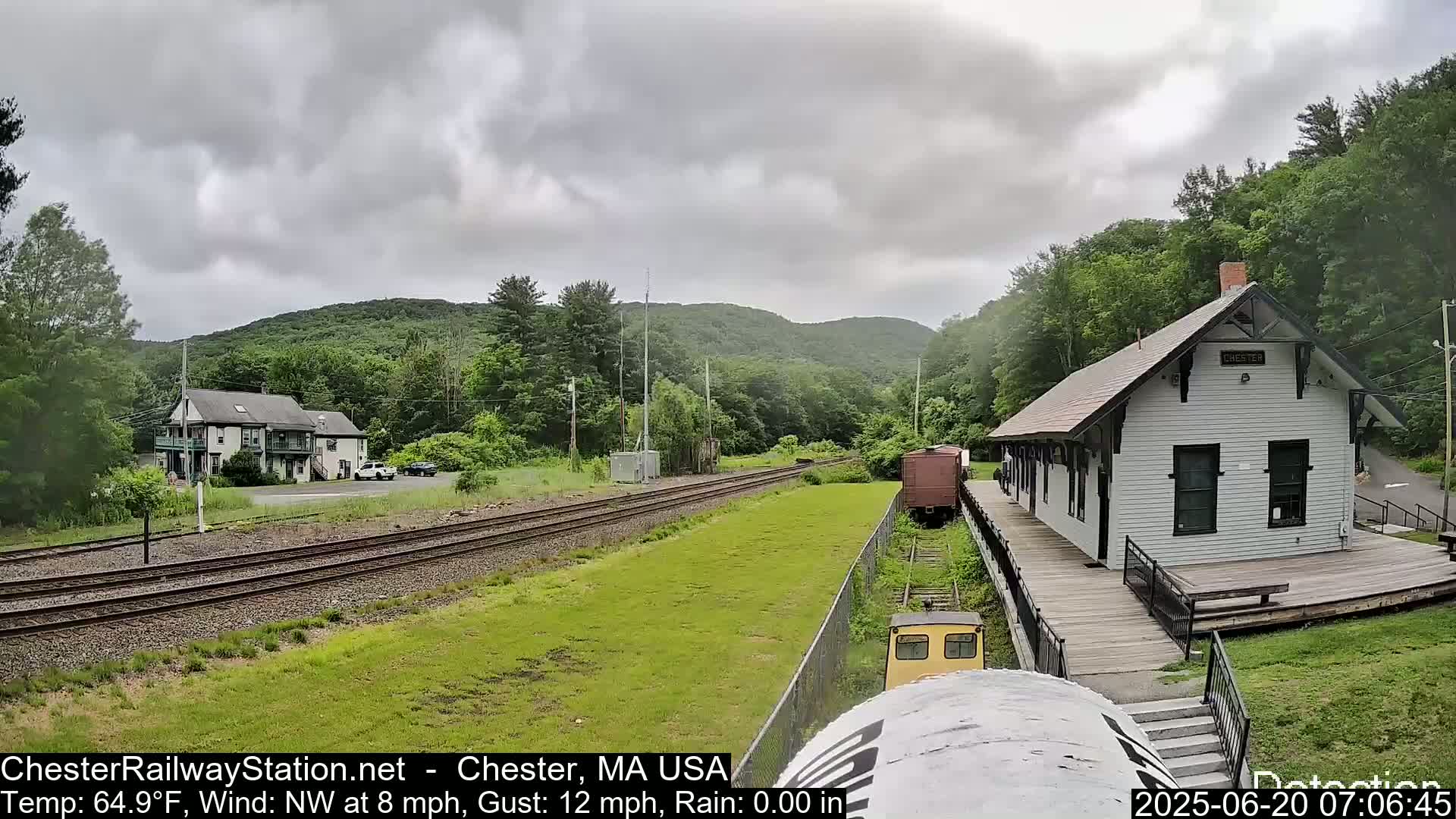 A view from a train shows a white train station, railroad tracks, a two-story building, and green hills under an overcast sky.