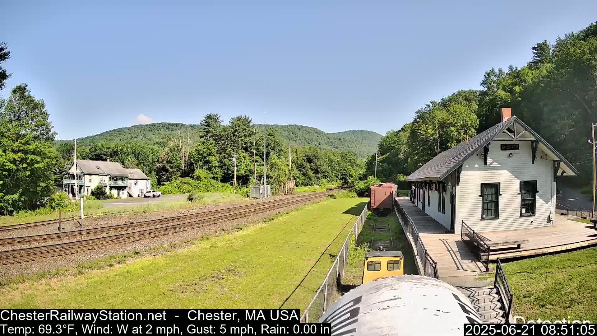 A white train station sits on a platform next to train tracks that lead toward a green, tree-covered mountain range under a clear sky.