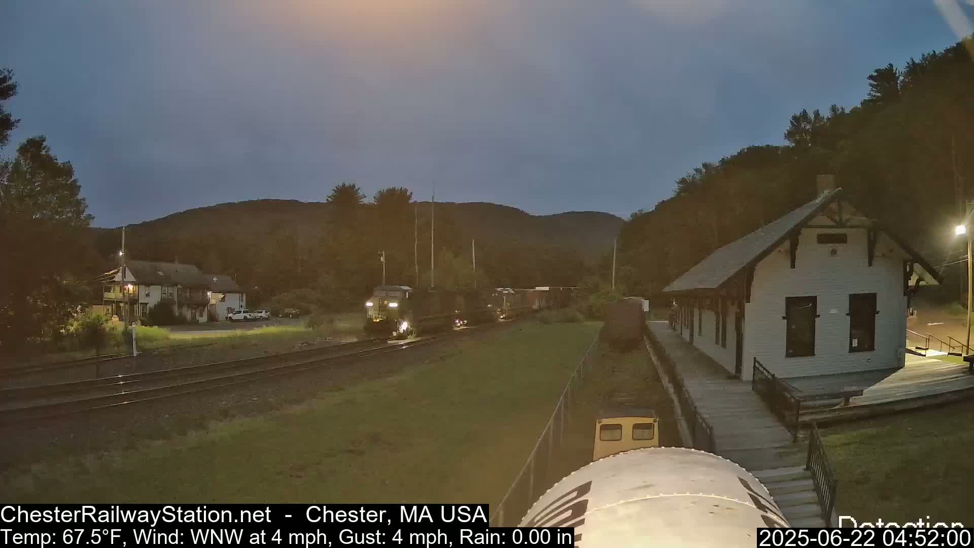 A freight train passes a train station under a twilight sky with a gentle wind.
