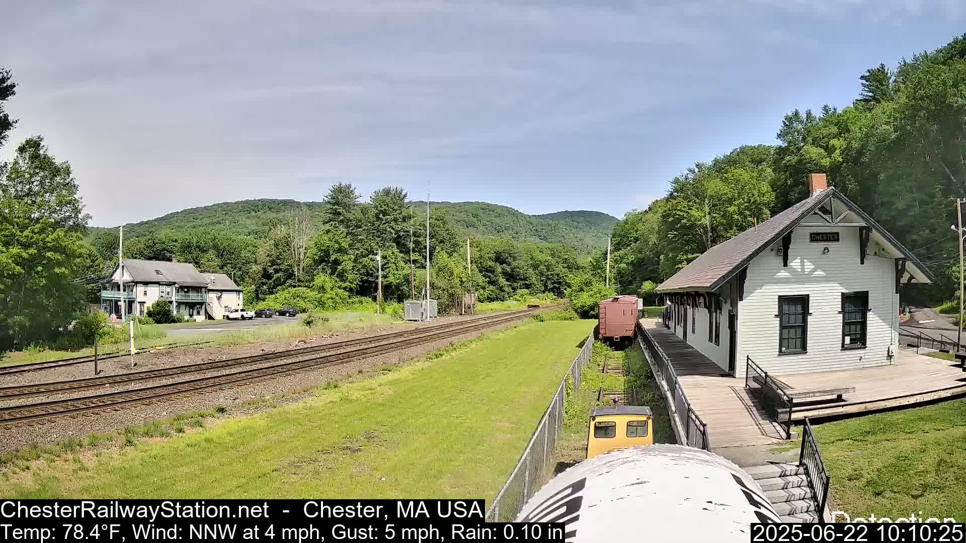 A white train station and a two-story building sit on either side of train tracks that stretch towards a backdrop of green hills under a partly cloudy sky.