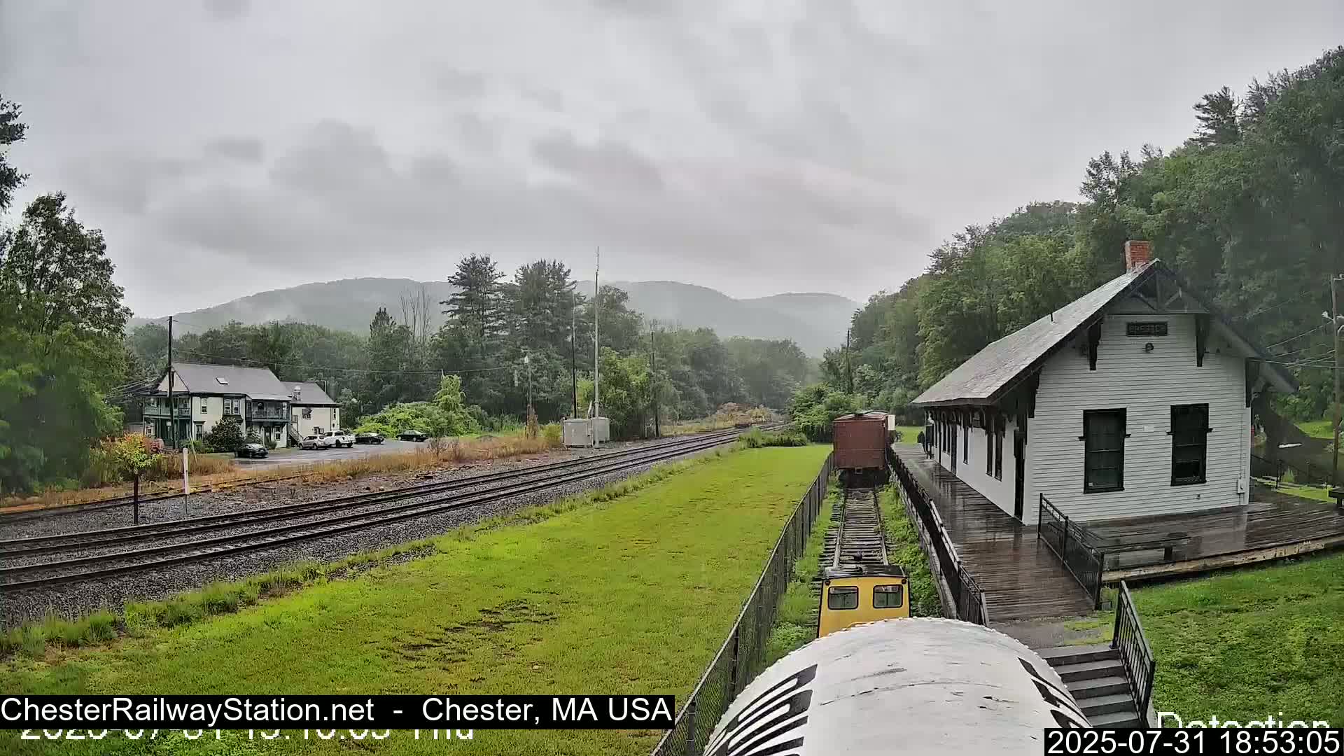 A small train approaches a white train station on a rainy day, with mountains and green trees in the background.