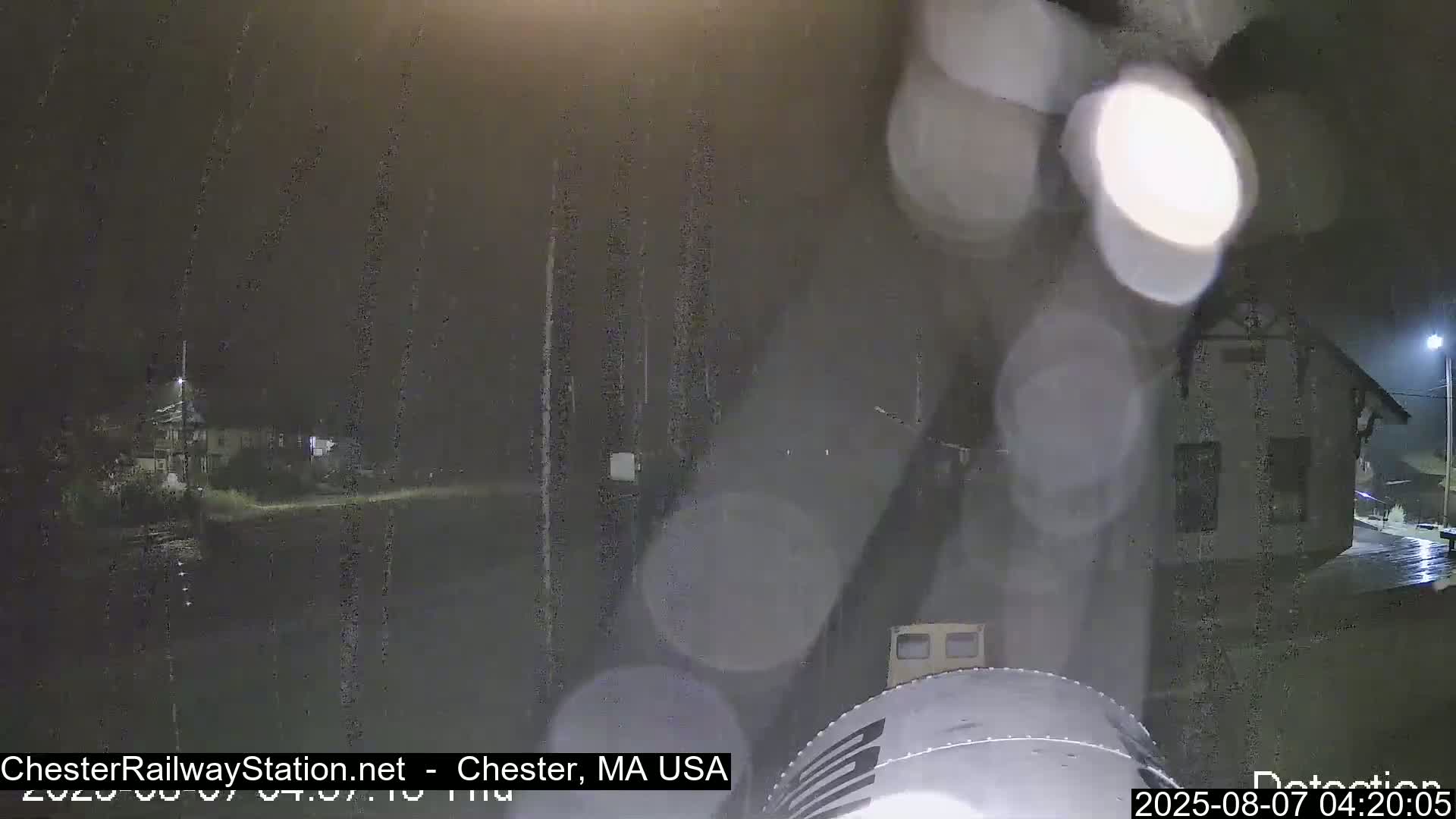A nighttime view of a train car in the rain, with a building visible in the background.