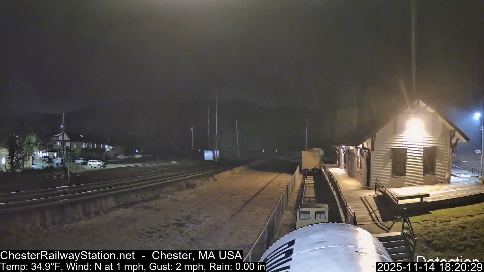 A clear night view depicts a dimly lit railway station with multiple tracks, an illuminated building and long platform, and several parked cars in the background under a dark sky.