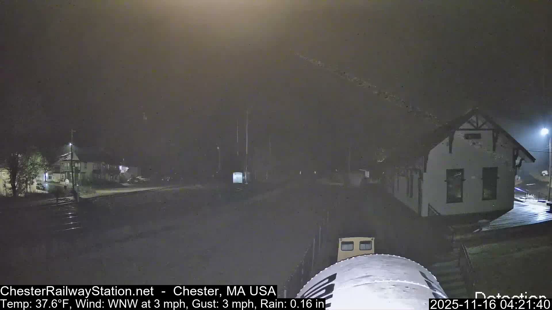 A foggy nighttime outdoor scene reveals diffused streetlights illuminating buildings, a street, and a partial view of a train on tracks.