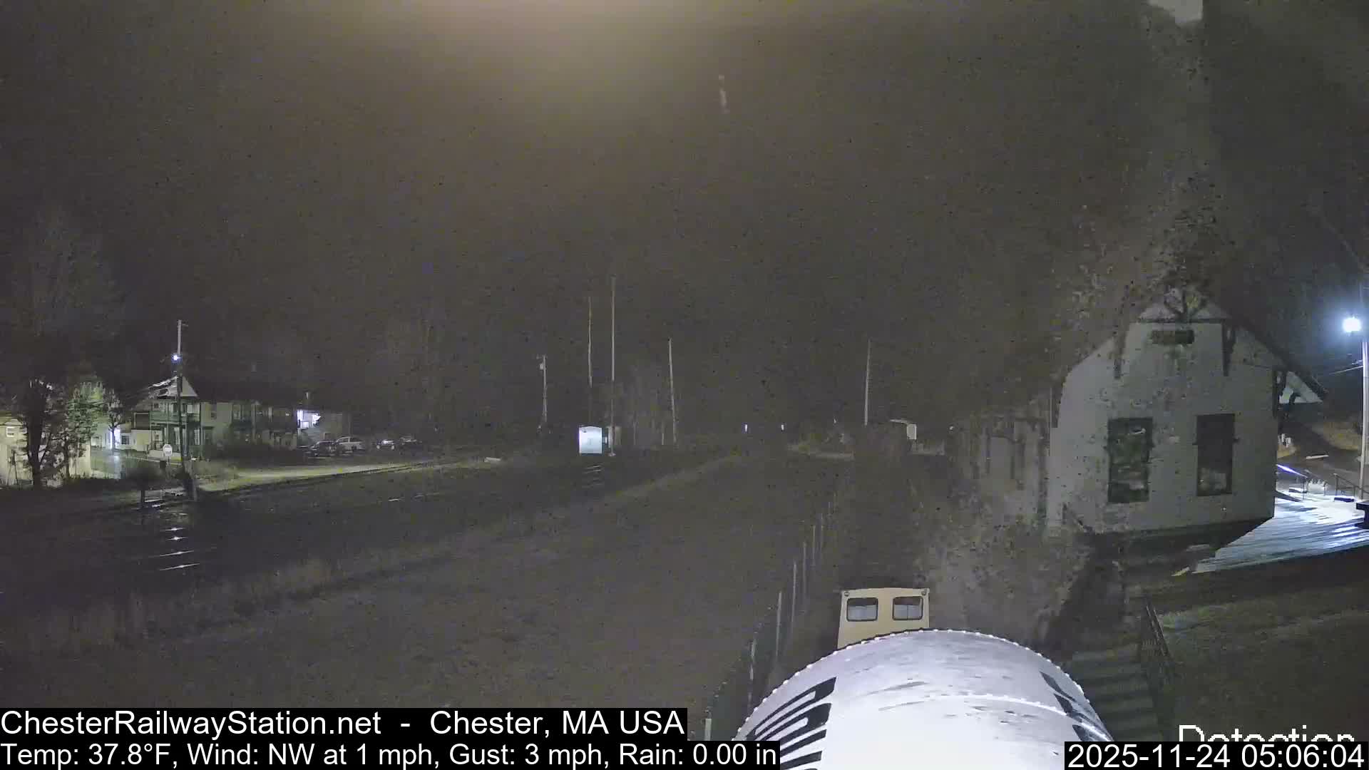 A grainy, low-light nighttime view reveals a railway track curving through a small town, flanked by residential buildings, bare trees, and an old station-like structure, with a large white object visible in the foreground, all under hazy conditions.