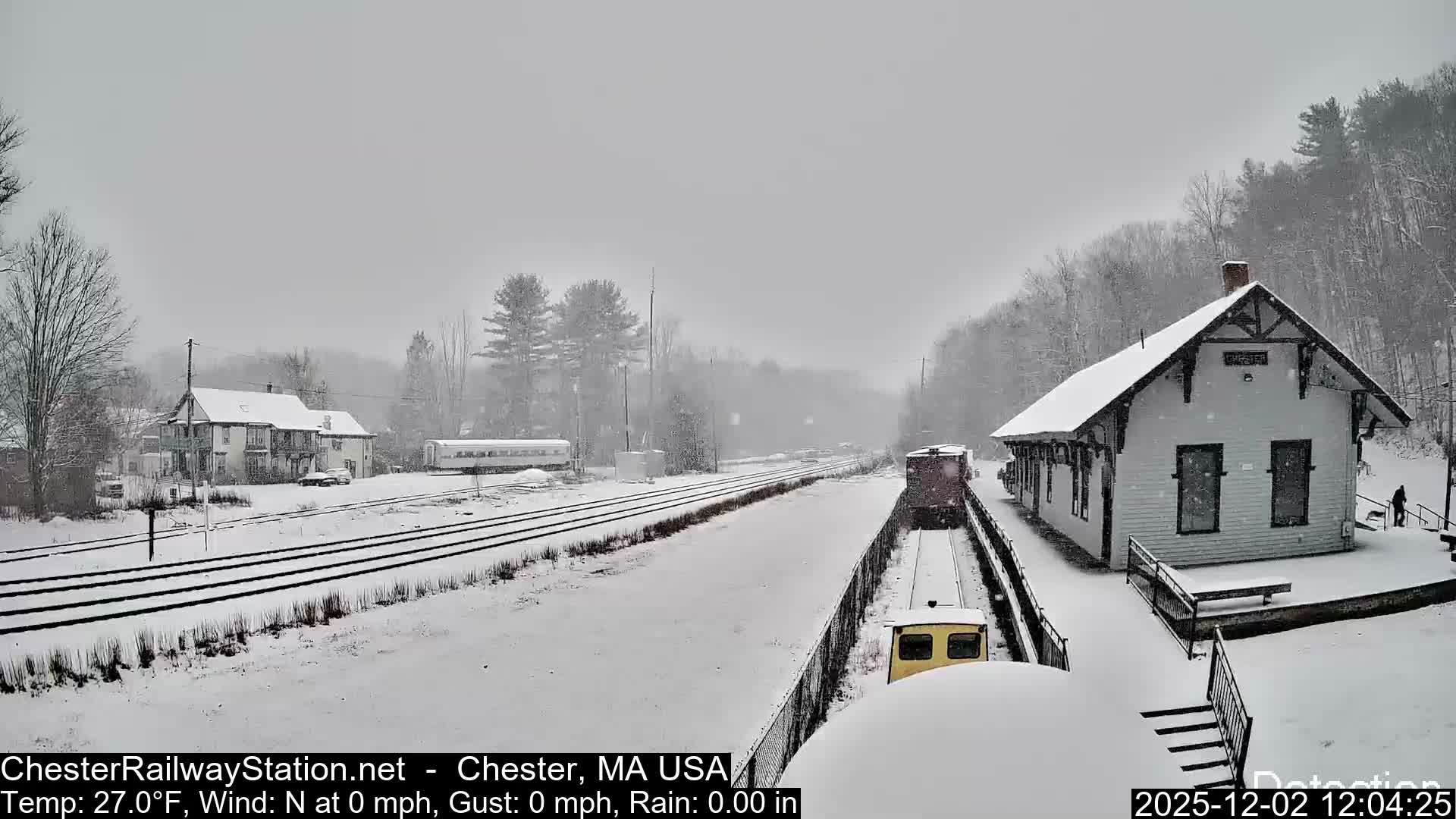 A snow-covered railway station featuring multiple tracks, several train cars, and a station building is depicted under a grey, overcast sky during a cold winter day.