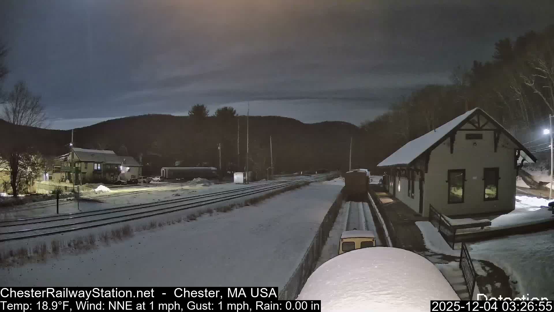 A snowy railway station scene at night features a well-lit building next to snow-covered tracks, with additional structures and distant hills visible under a dark, cloudy sky.