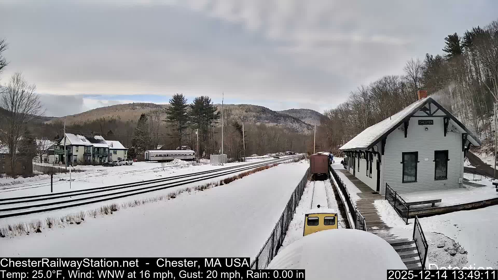 A snowy railway station scene at night features a well-lit building next to snow-covered tracks, with additional structures and distant hills visible under a dark, cloudy sky.