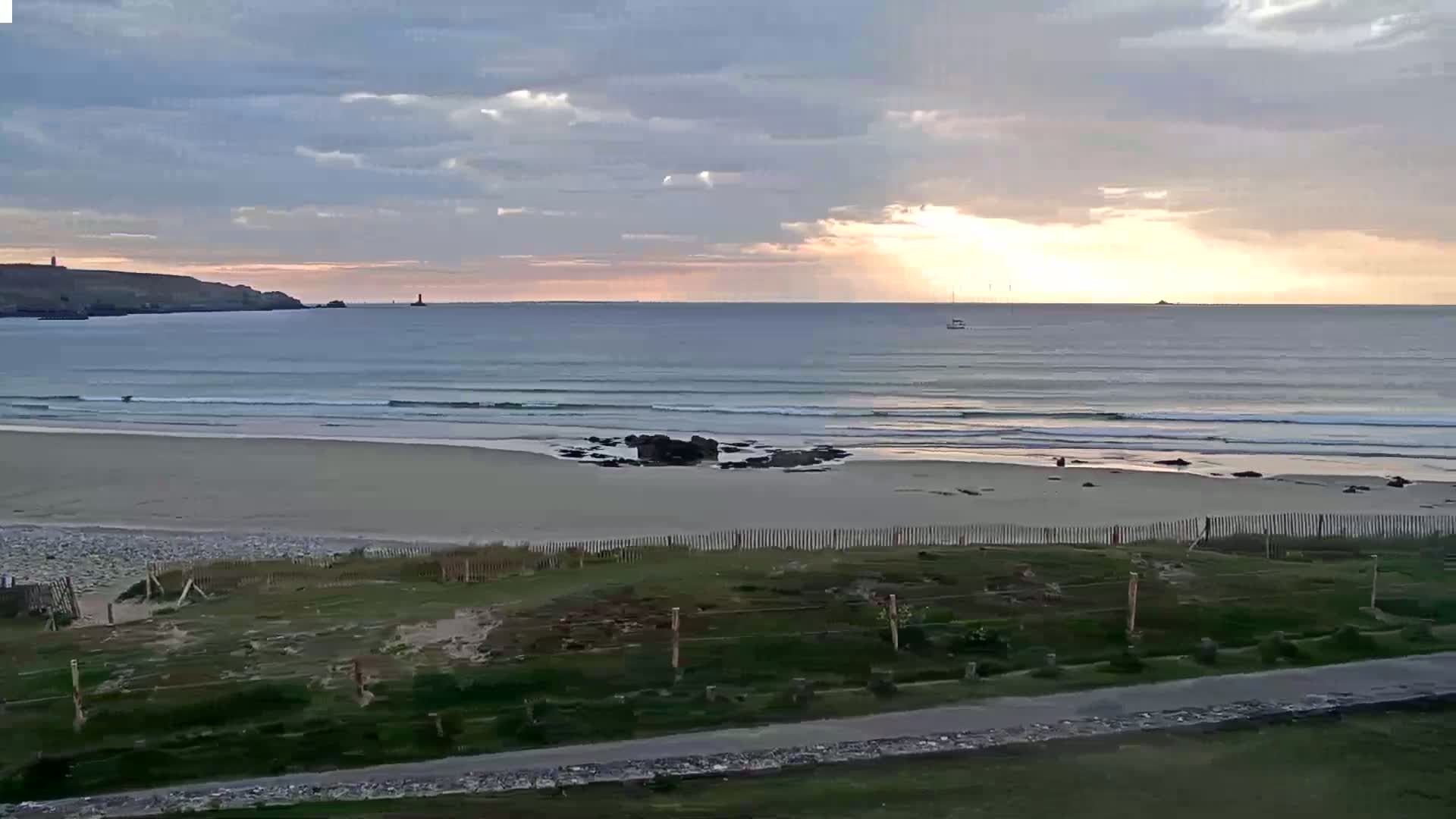 A sandy beach with gentle waves rolls onto the shore under a partly cloudy sunrise sky.