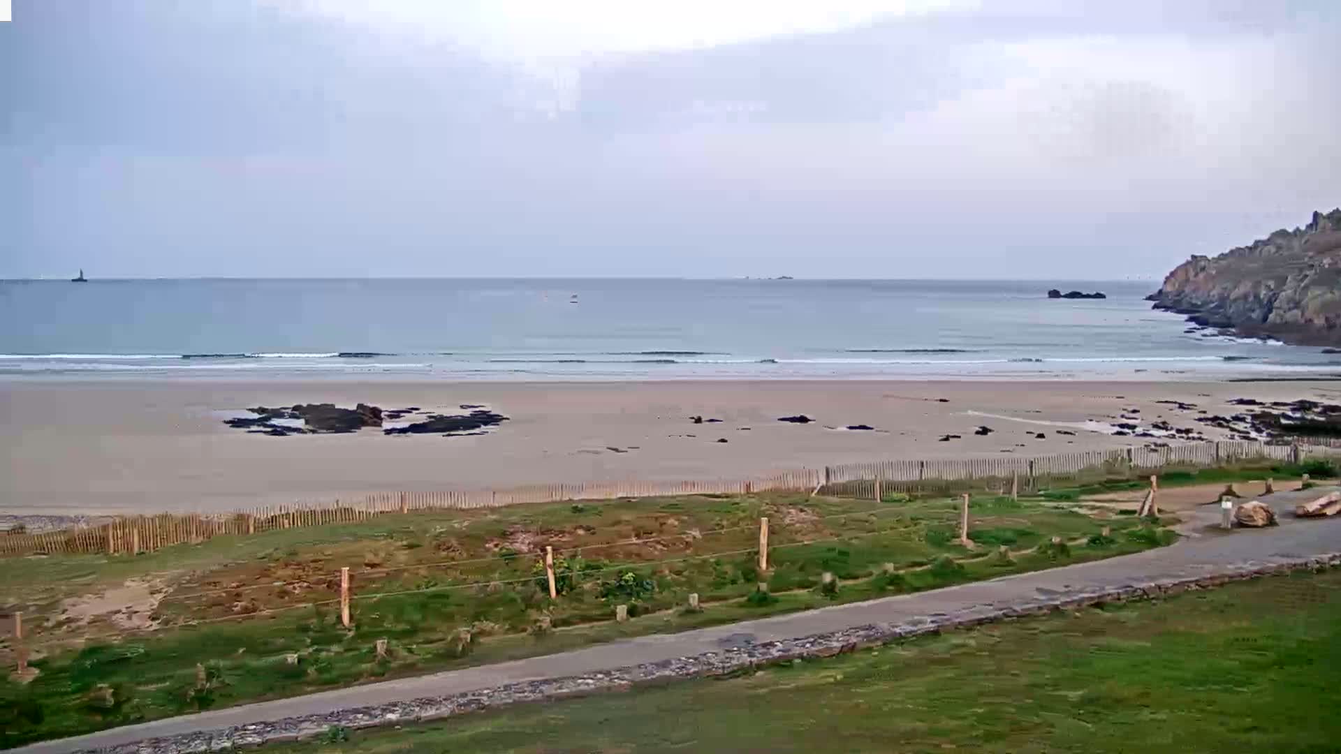 A sandy beach with small waves stretches to a calm ocean under an overcast sky, bordered by grassy land and a low stone wall.