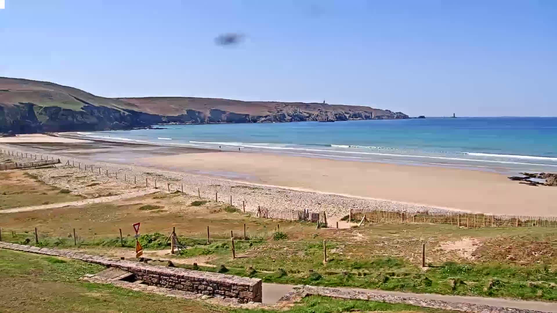A sandy beach with small waves laps against the shore under a clear blue sky, backed by grassy hills and a distant lighthouse.