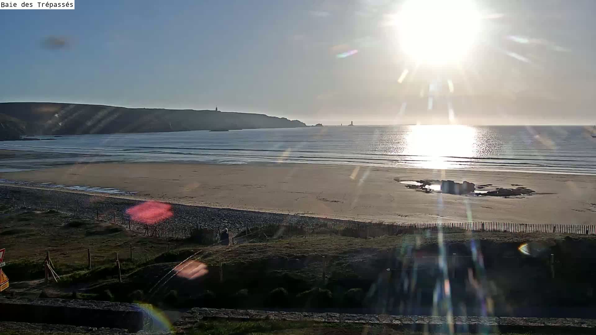A sandy beach with calm ocean waves stretches out under a bright, sunny sky, backed by dark cliffs and distant lighthouses.