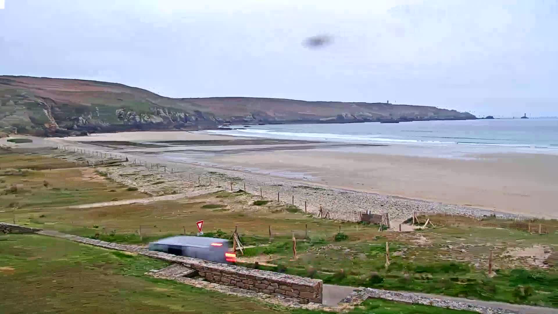 A gray vehicle drives along a road overlooking a long sandy beach with gentle waves under an overcast sky.