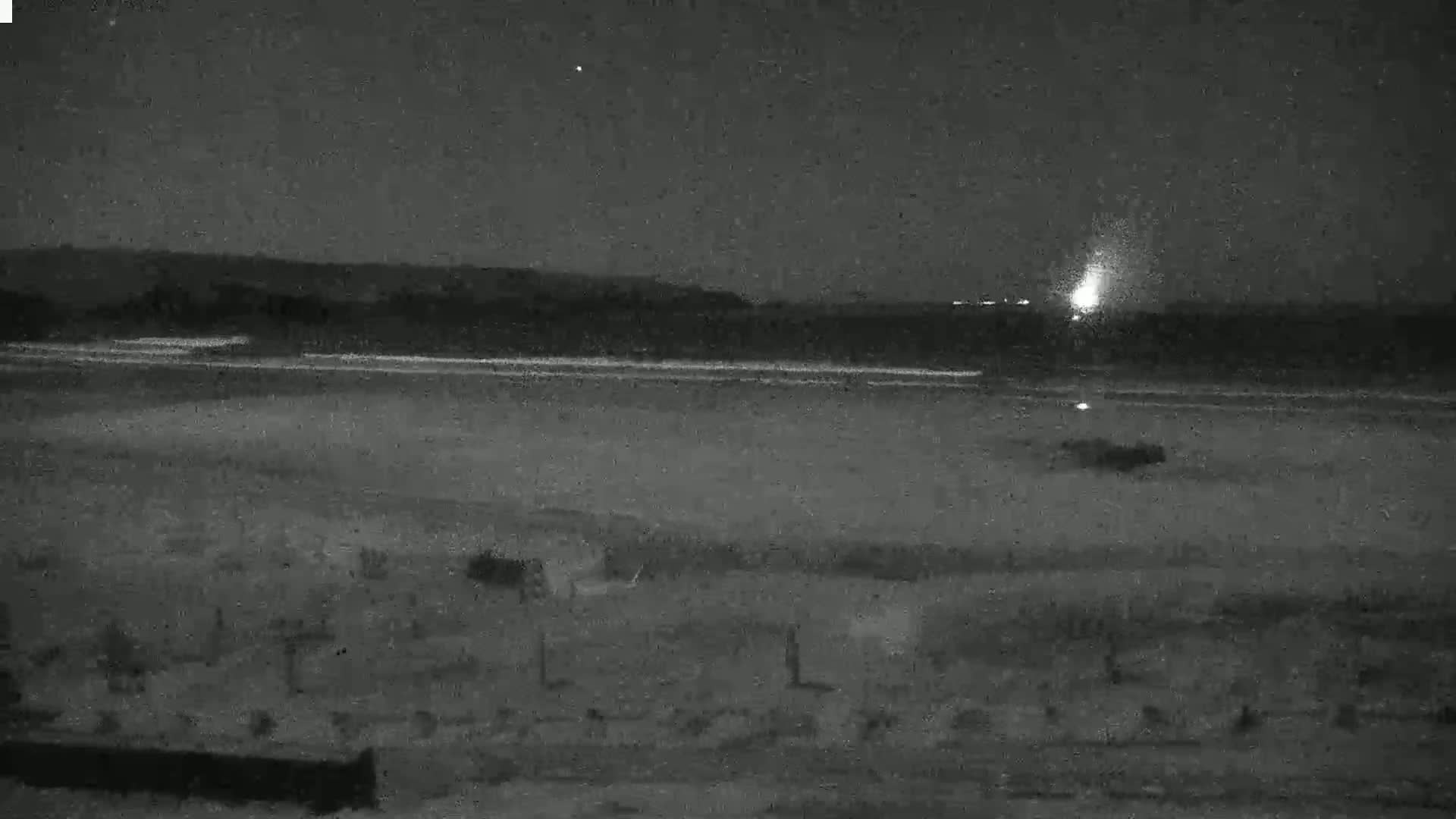 A nighttime, low-light view shows a dark beach with a distant, bright light source over a body of water, under a clear sky.