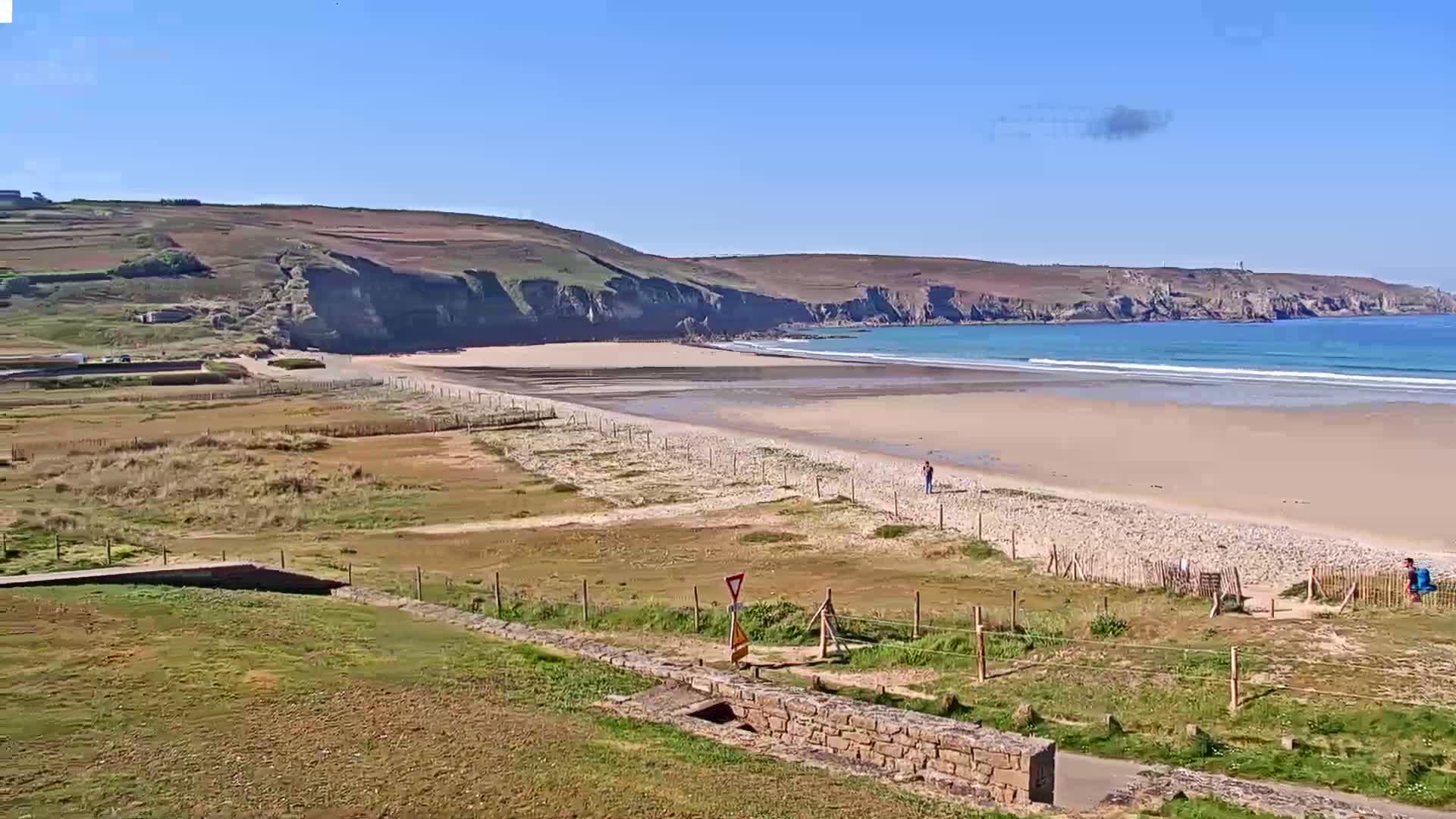 A long, sandy beach stretches below a rocky cliff face under a clear, sunny sky, with a few people visible on the beach and a grassy area in the foreground.