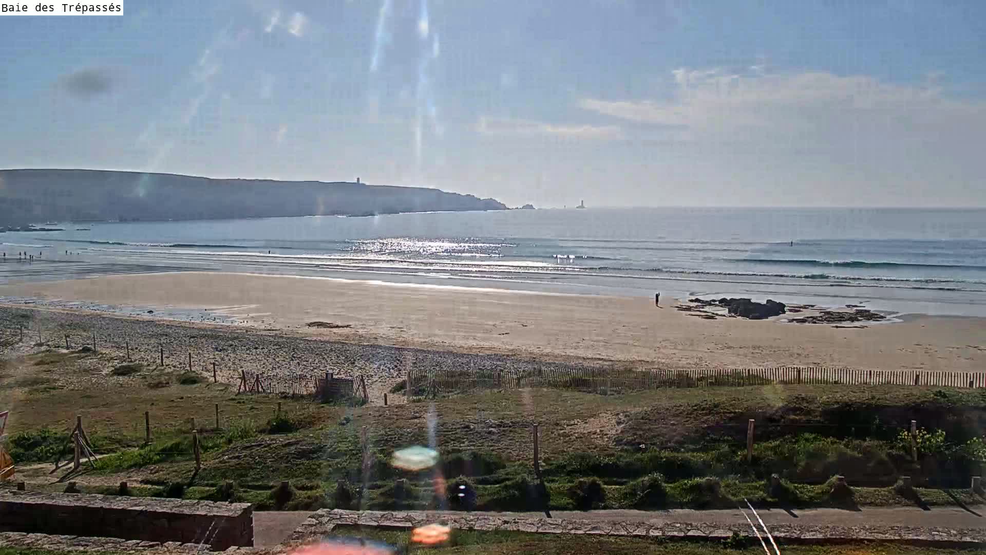 A sandy beach with small waves and a few people stretches out to meet a calm ocean under a mostly sunny sky.