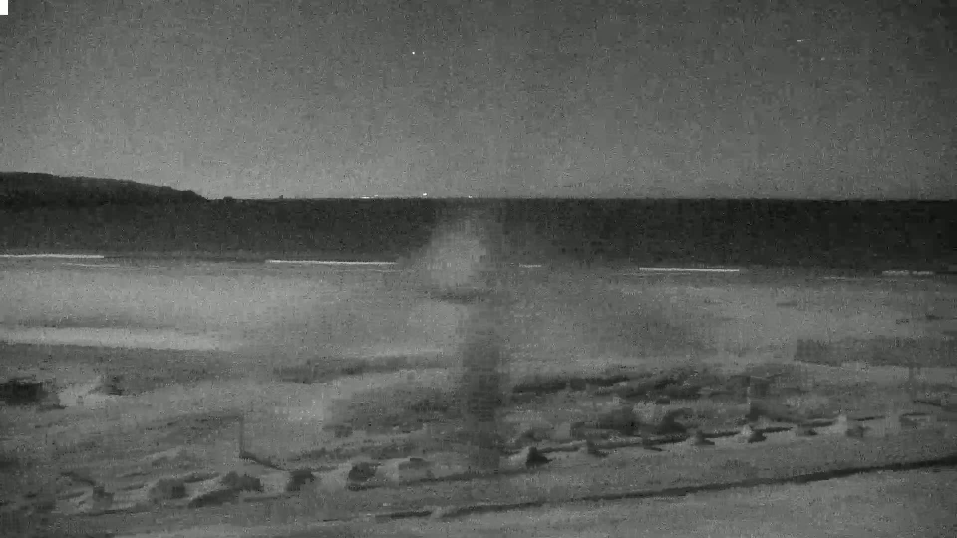 A nighttime, low-light view shows a dark beach with a body of water and a distant shoreline under a dark sky.