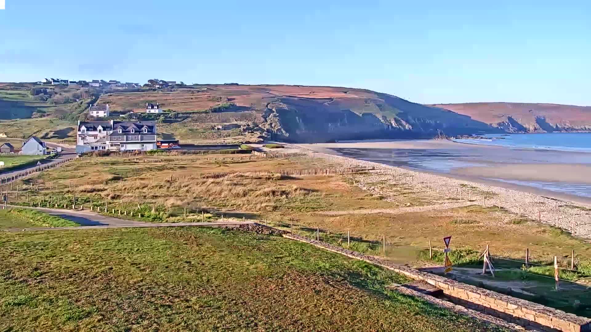 A long sandy beach stretches out below a coastal village nestled against a hillside under a clear, sunny sky.