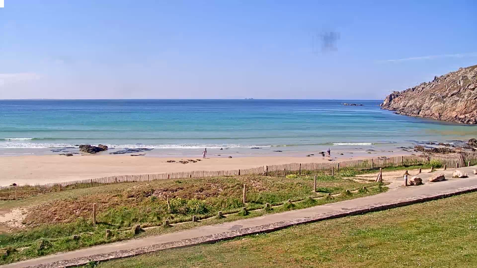 A sandy beach with small waves and a few people on it is bordered by a wooden fence and grassy area, all under a clear blue sky.