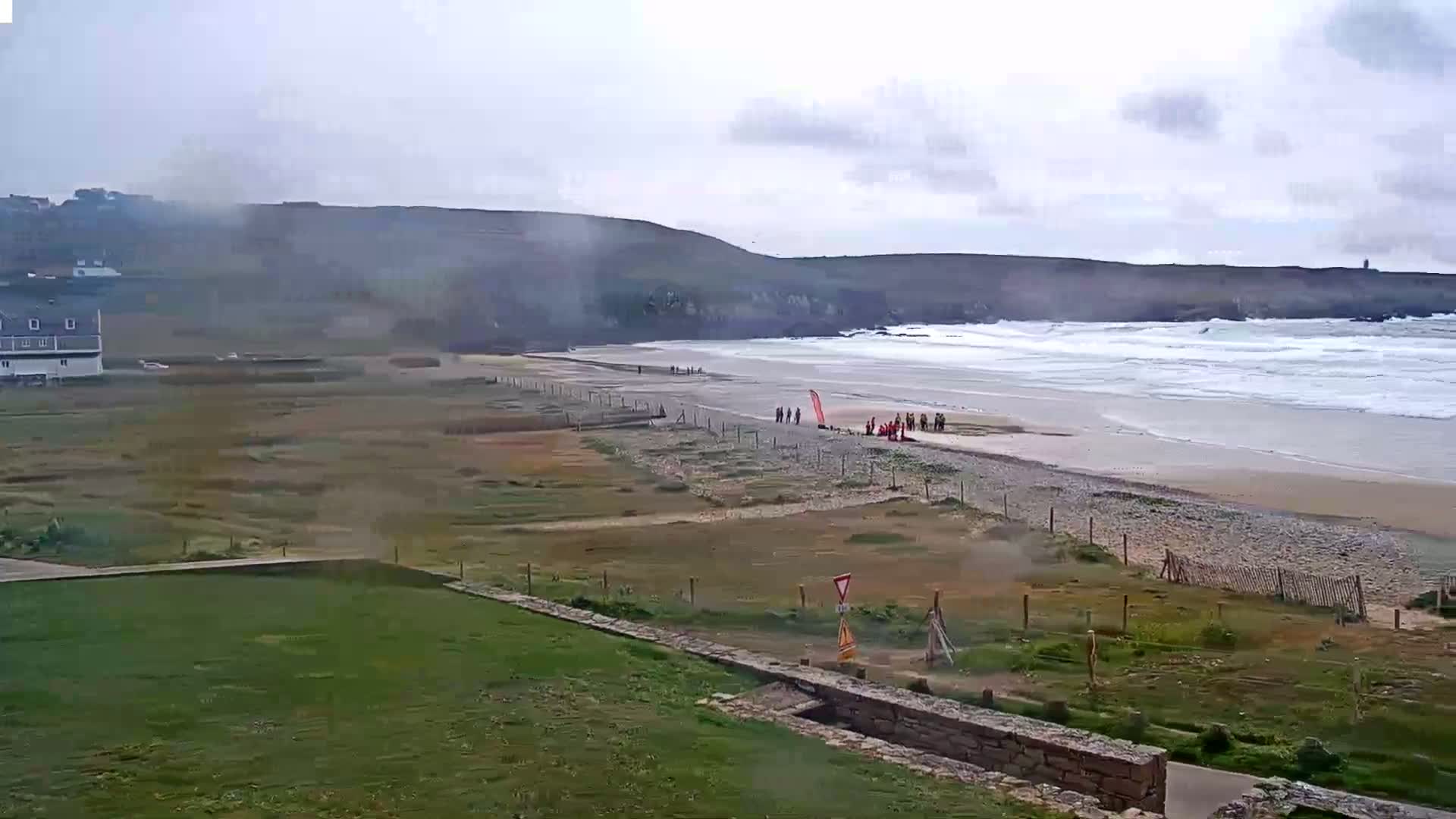 A wide shot shows a beach with moderate waves under an overcast sky, a group of people standing near the water's edge, and a building in the distance.