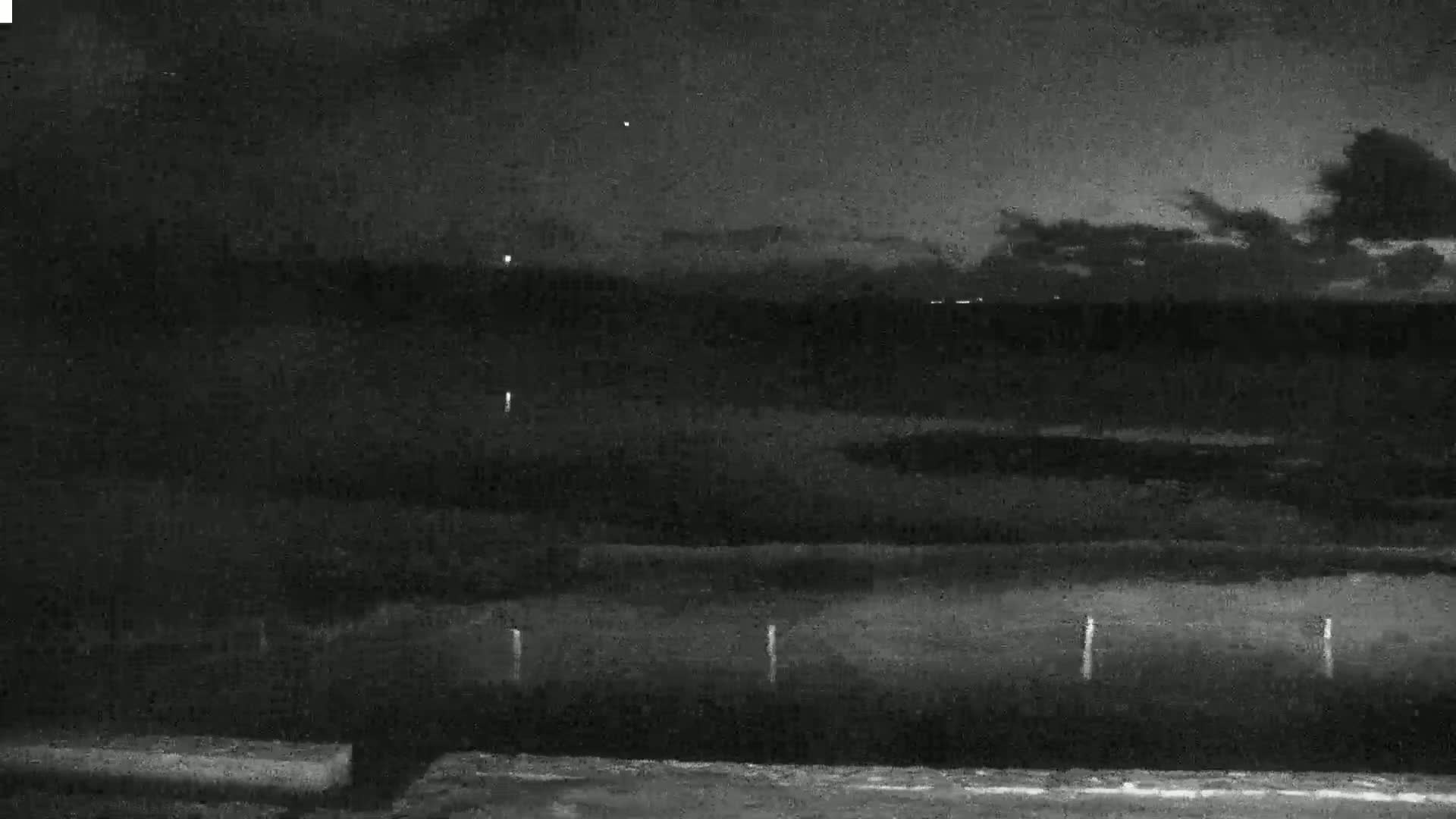 A nighttime, overcast view of a dark landscape with a few distant lights and several thin, vertical posts in the foreground.