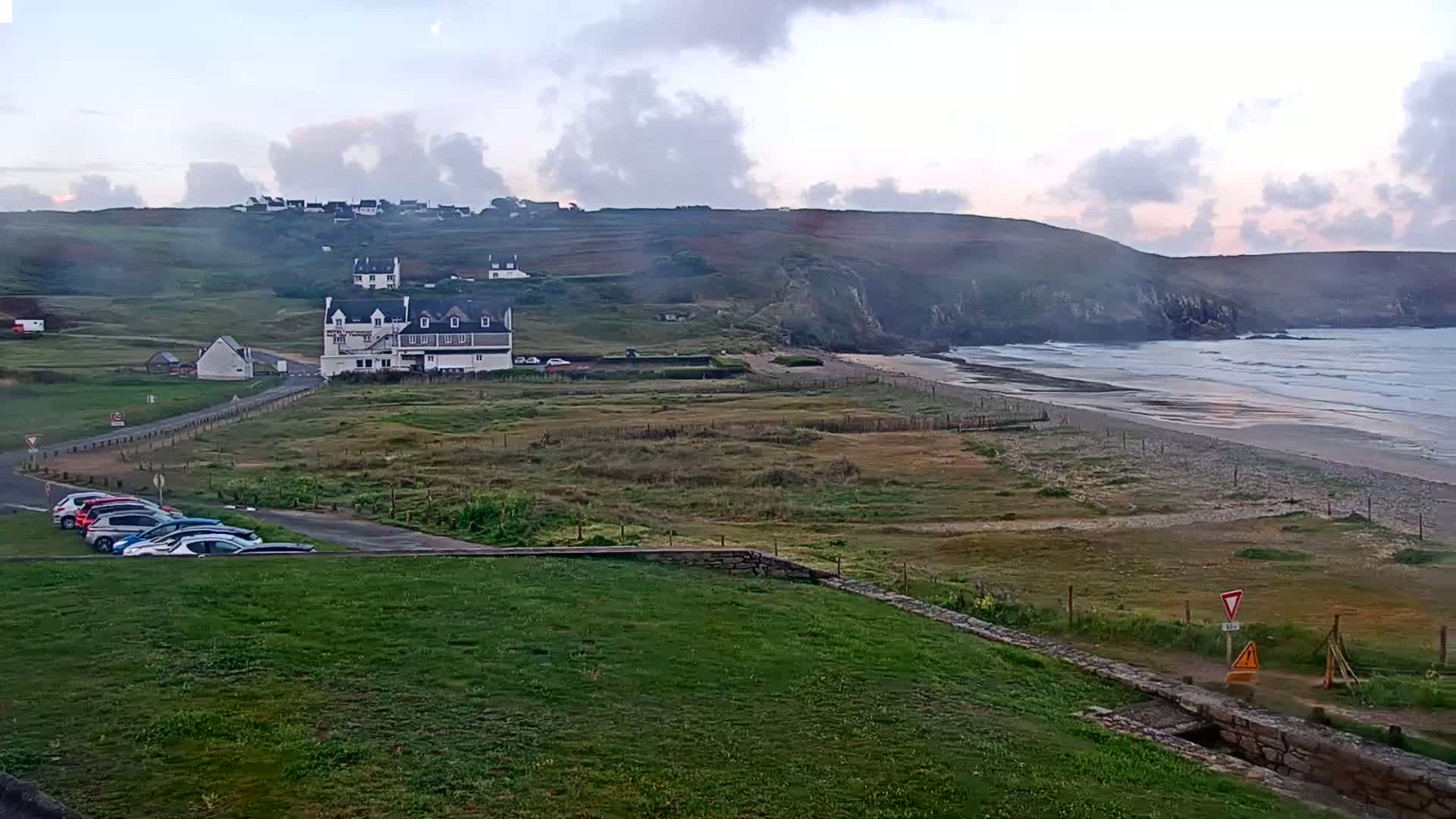 A coastal scene under a cloudy sky shows a beach with gentle waves, a grassy area with a parking lot and several buildings, including a large white hotel, situated on a hillside overlooking the ocean.