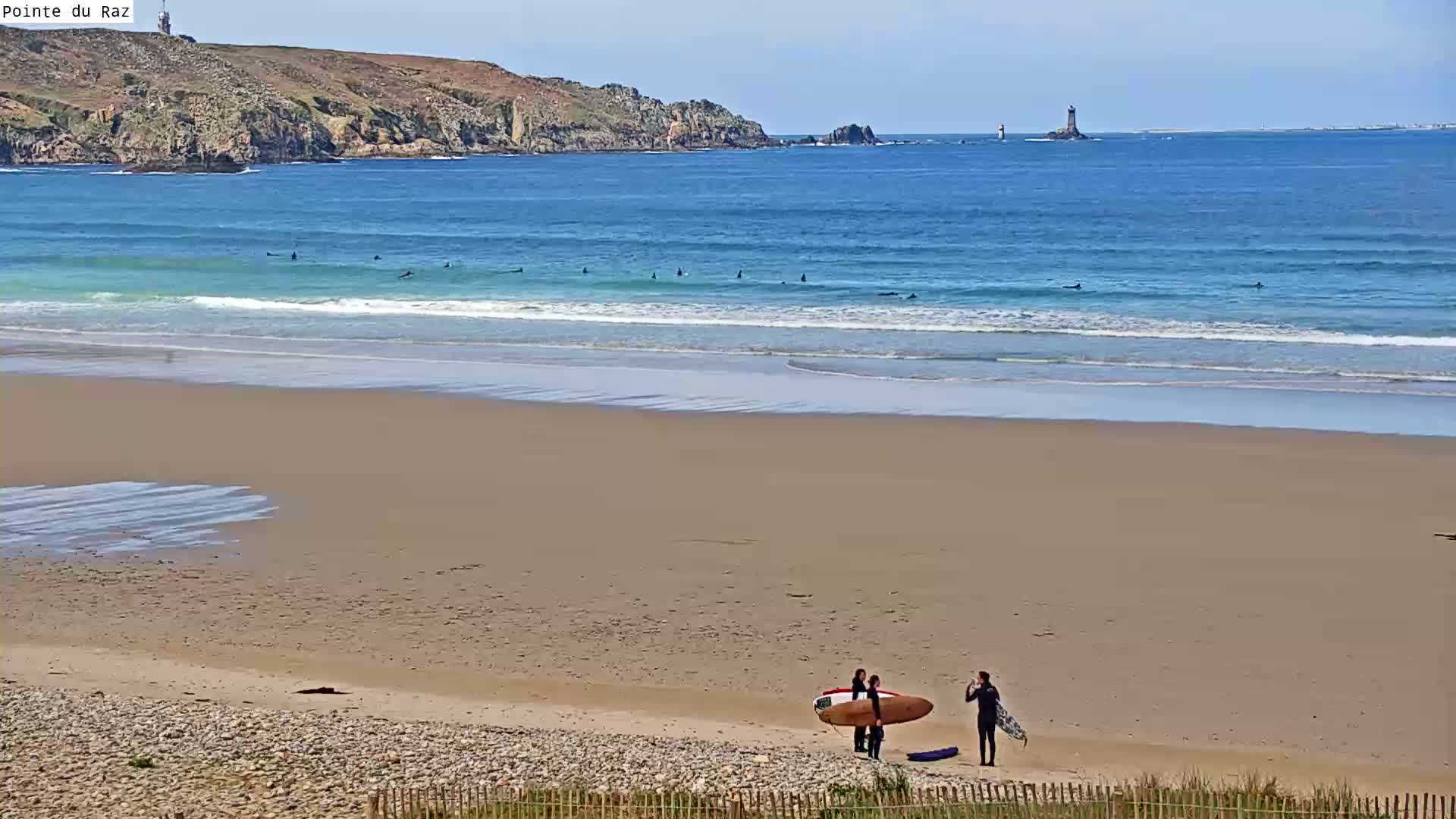 A sandy beach with several surfers in the ocean and three people standing near surfboards on the shore under a clear sky.