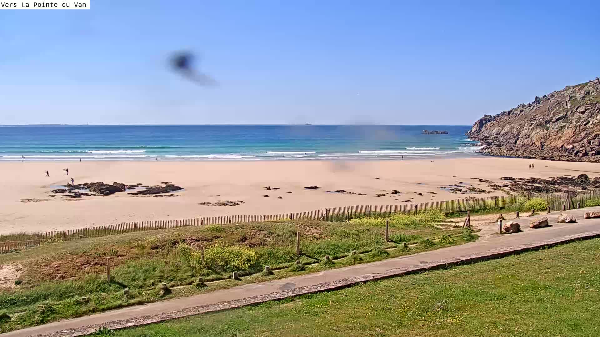 A sandy beach with gentle waves under a clear blue sky is enclosed by a wooden fence and overlooks a grassy area with a paved path.