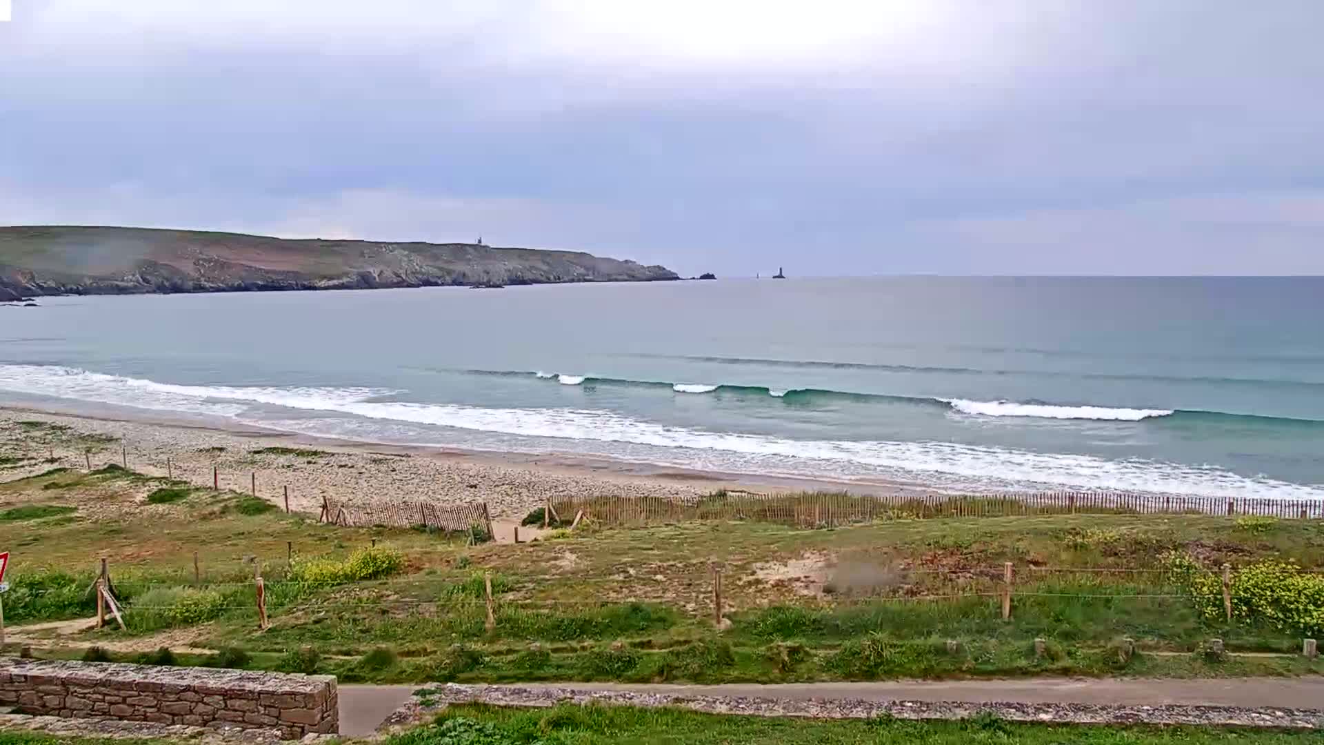Under a cloudy sky, gentle waves roll onto a sandy beach backed by grassy dunes and a low stone wall.