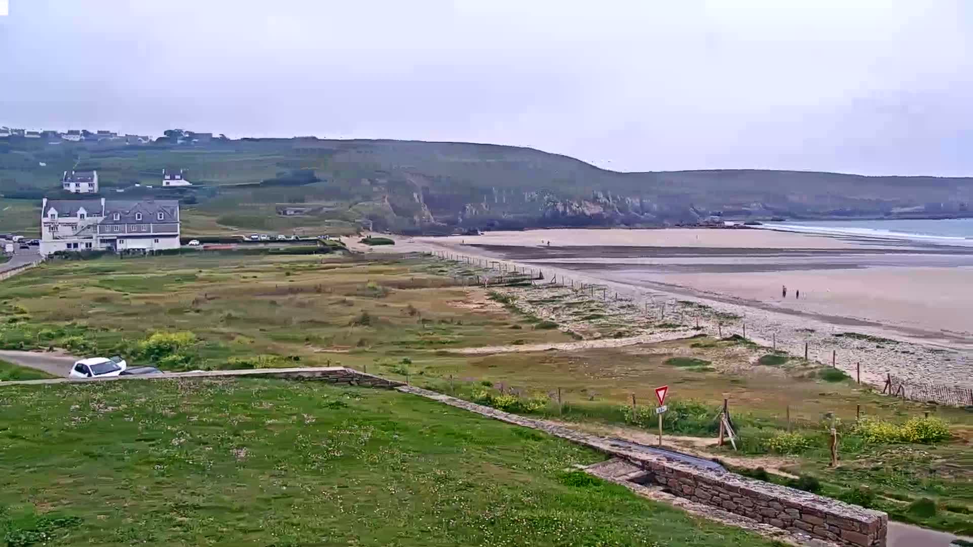 A sandy beach stretches below a grassy hillside, featuring a building and some cars near the top, under an overcast sky.