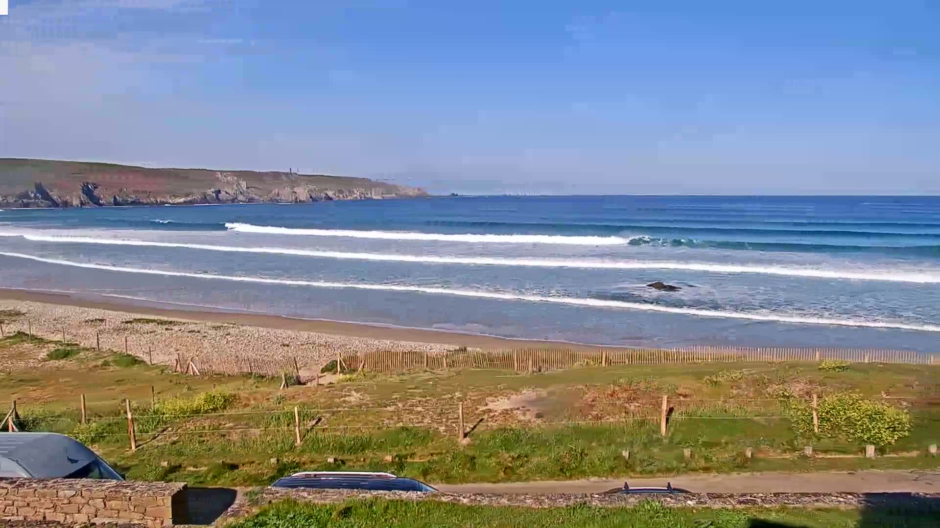 Under a clear blue sky, gentle waves roll onto a sandy beach bordered by grassy dunes and a low stone wall.