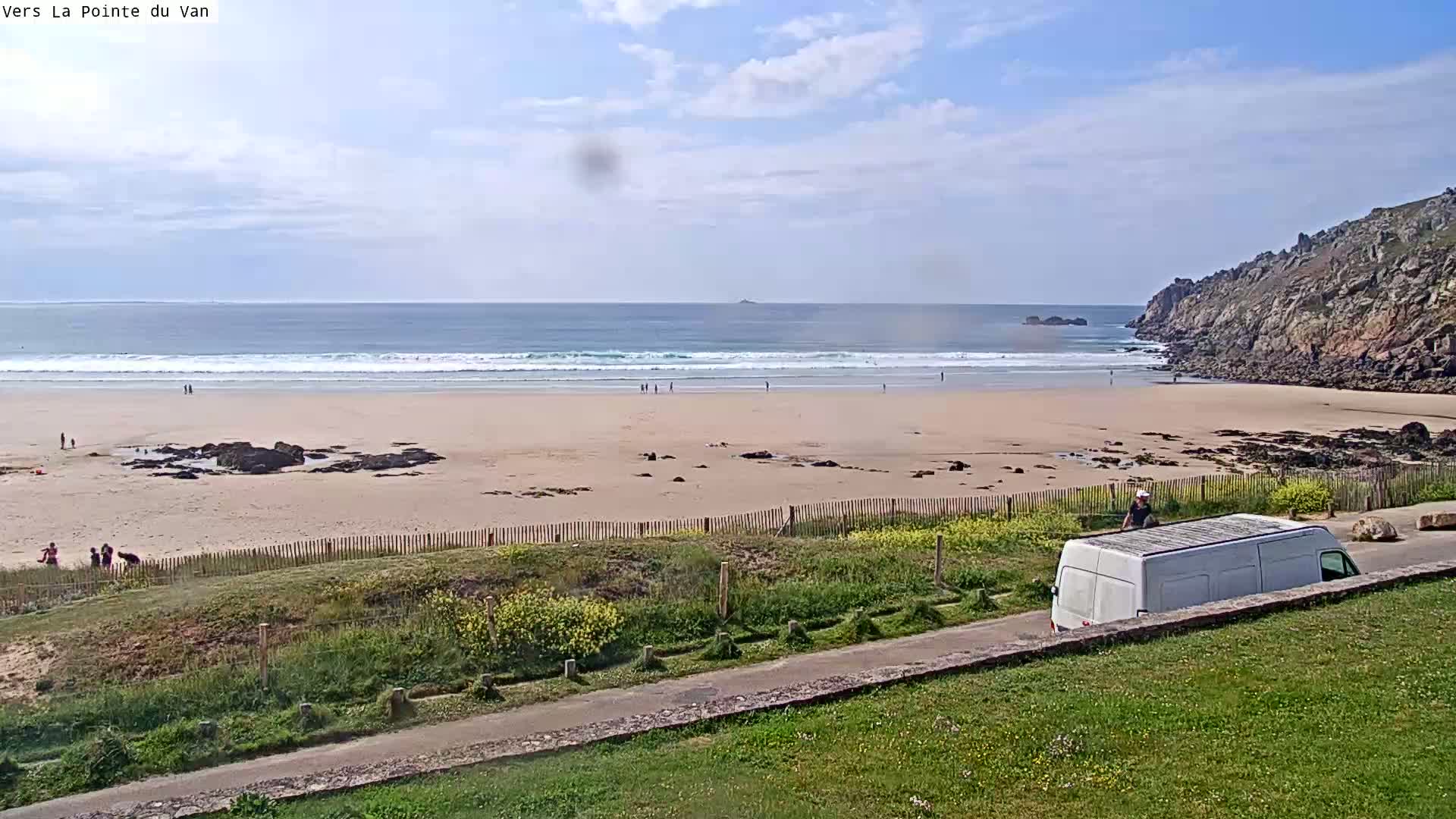 A sandy beach with small waves and a few people is partially enclosed by rocky cliffs under a partly cloudy sky, and a white van is parked near a road overlooking the scene.