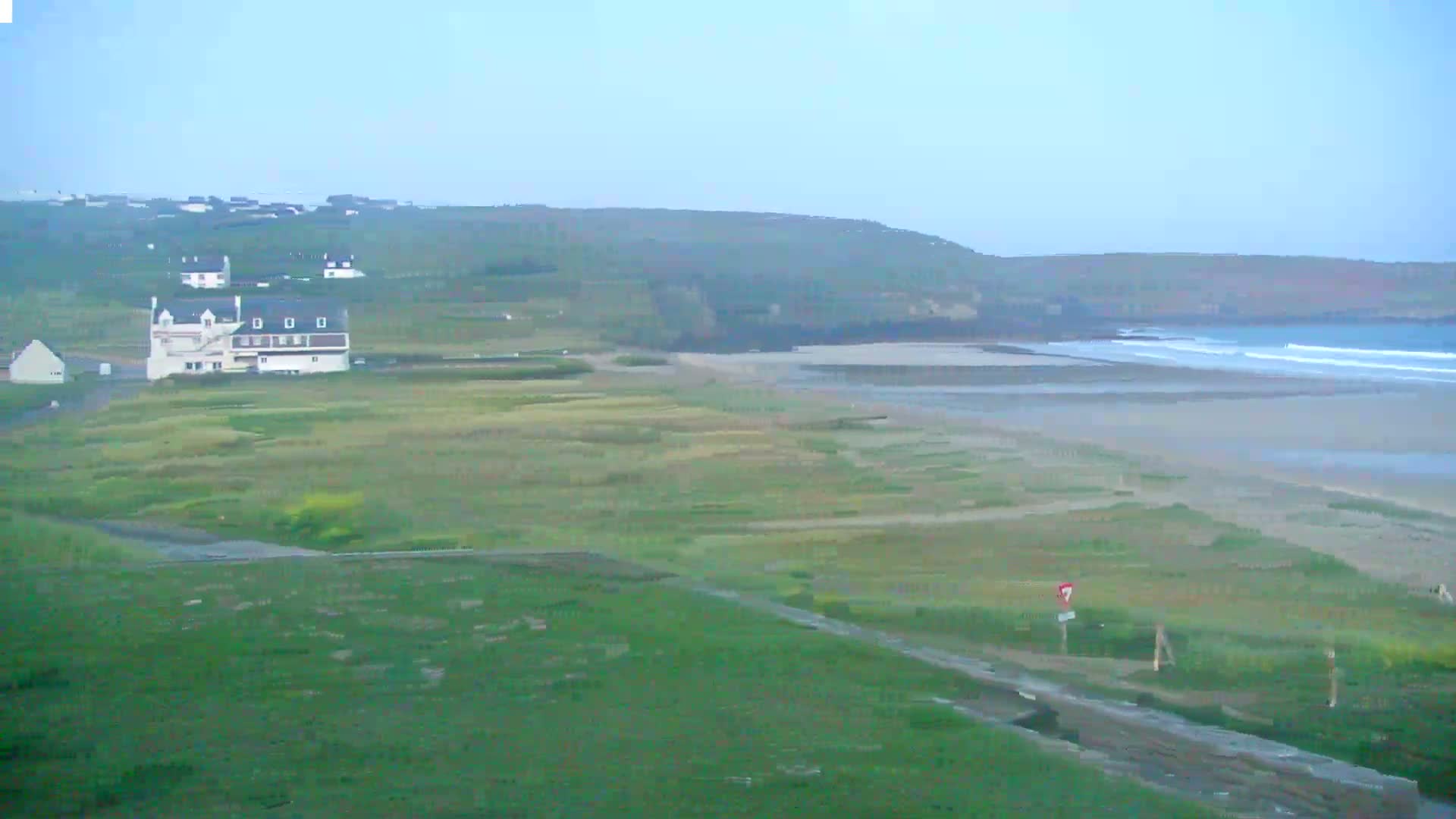 A white building sits near a sandy beach with small waves under a hazy sky.