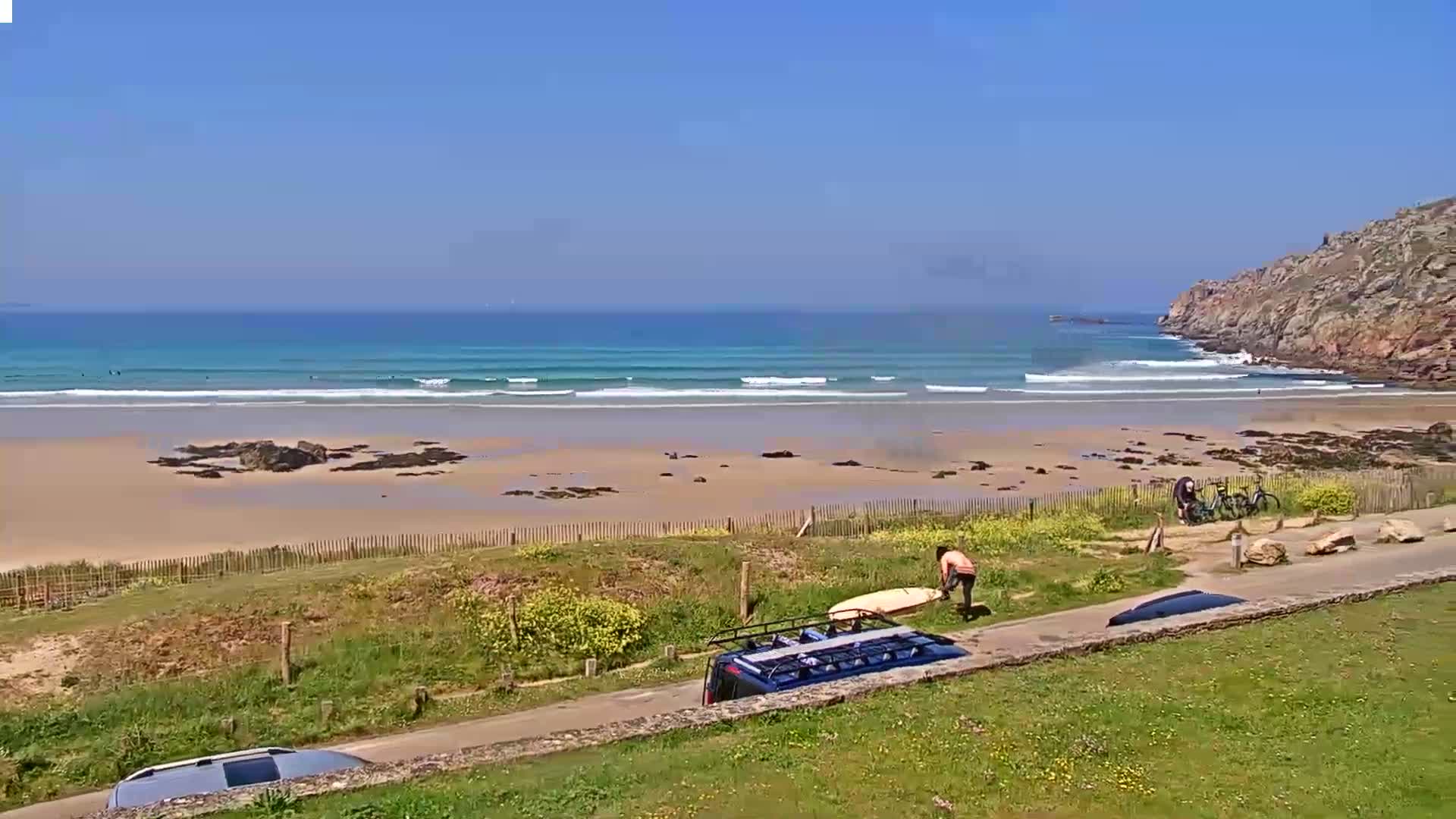 A sandy beach with small waves under a clear blue sky is viewed from a grassy hillside, where several people are near vehicles and bicycles.