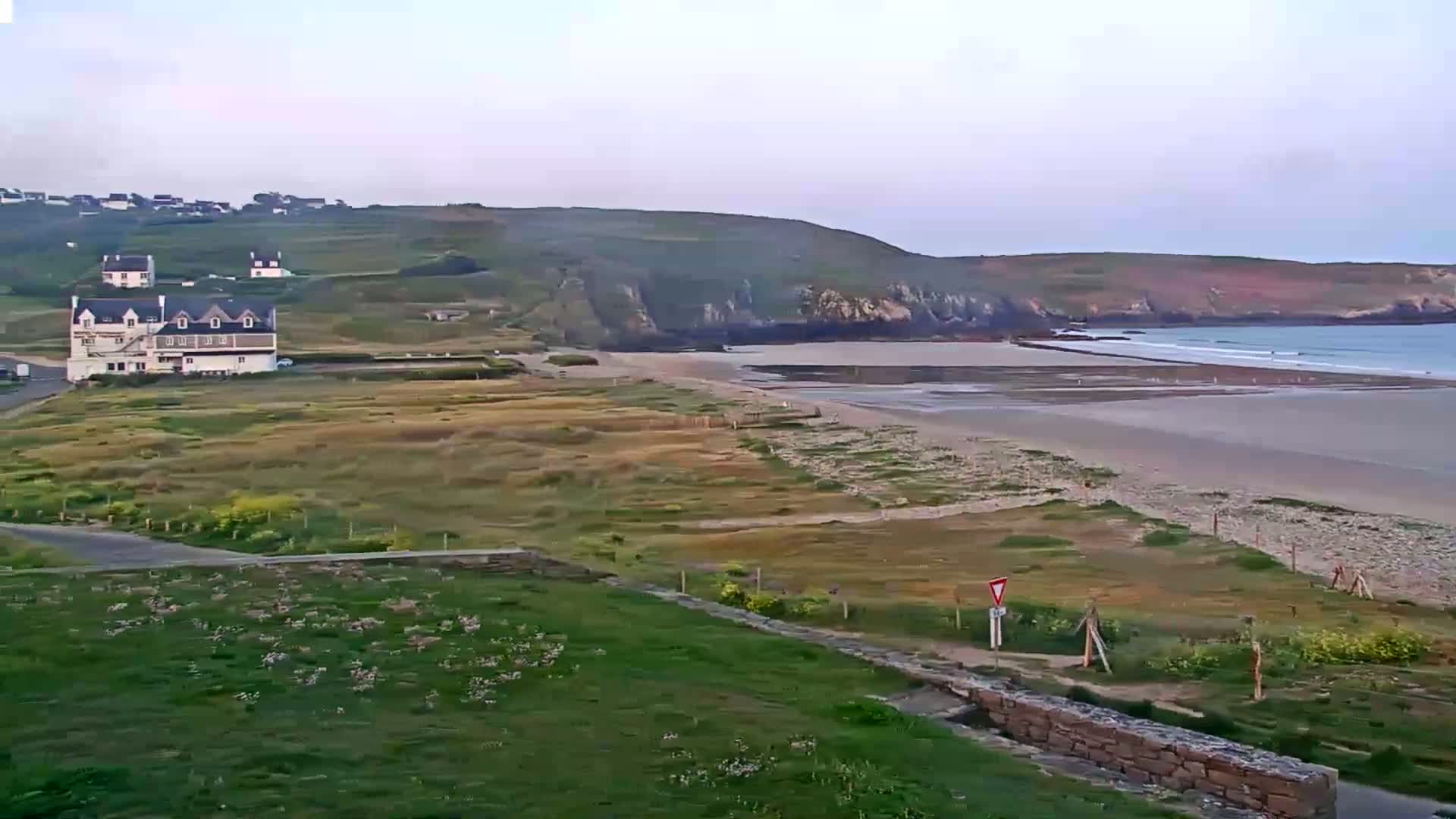 A sandy beach stretches below a grassy hillside with buildings and a small town in the distance under a mostly clear sky.
