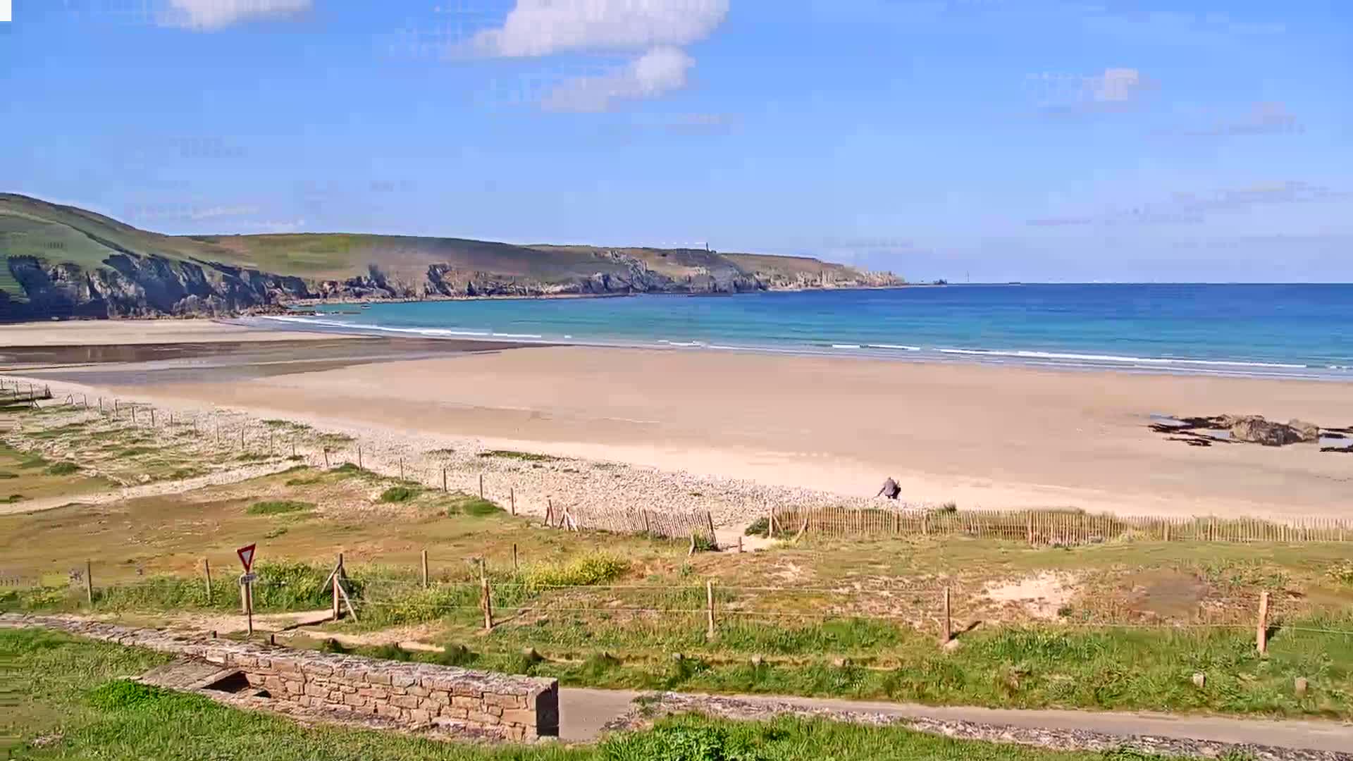 A sandy beach stretches out to a calm, turquoise ocean under a mostly sunny sky, backed by grassy hills.