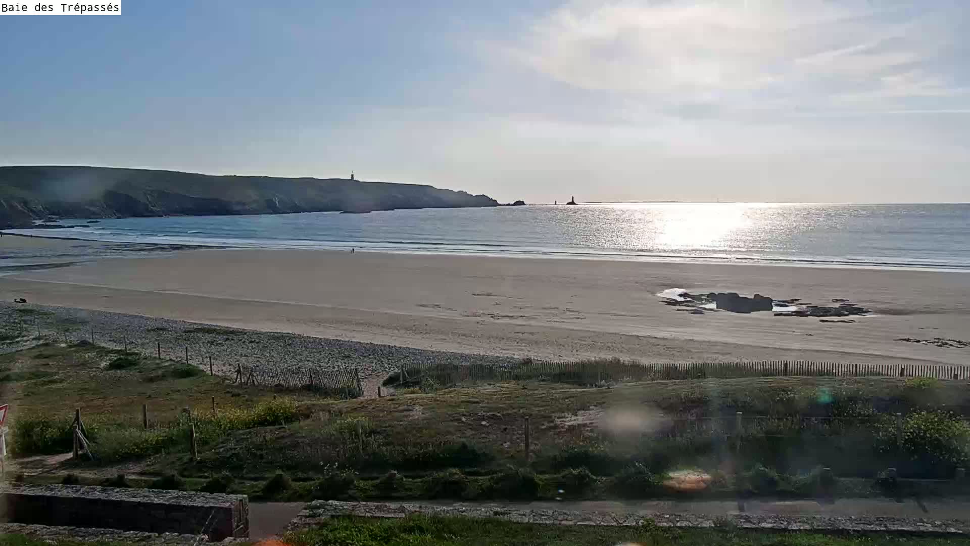 A sandy beach stretches out to a calm ocean under a mostly sunny sky, with a distant coastline and a lighthouse visible.
