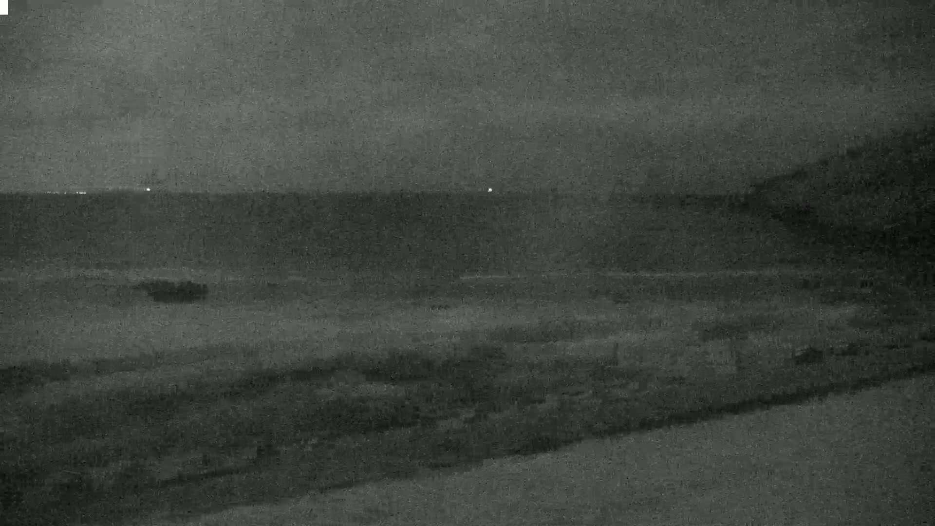 A low-light, nighttime view shows dark land sloping gently down to a dark body of water under a cloudy sky.