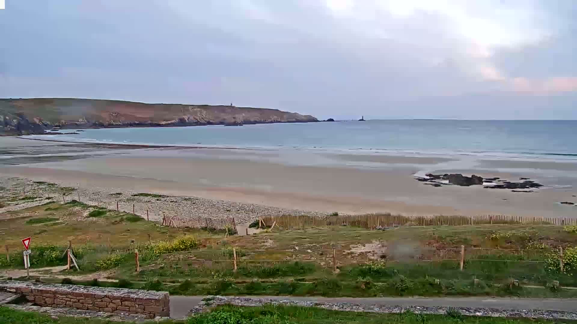 A sandy beach stretches out to a calm, light blue ocean under an overcast sky, backed by a rocky headland.