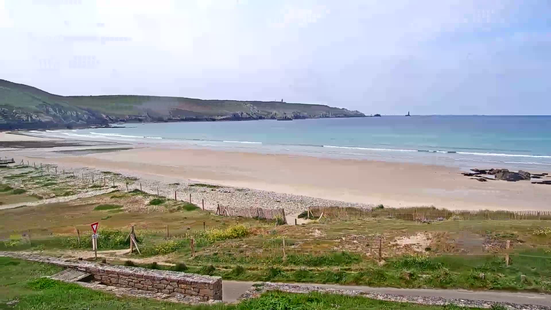 A long sandy beach stretches out to a calm, light blue ocean under a mostly cloudy sky, with green hills and grassy dunes in the foreground.