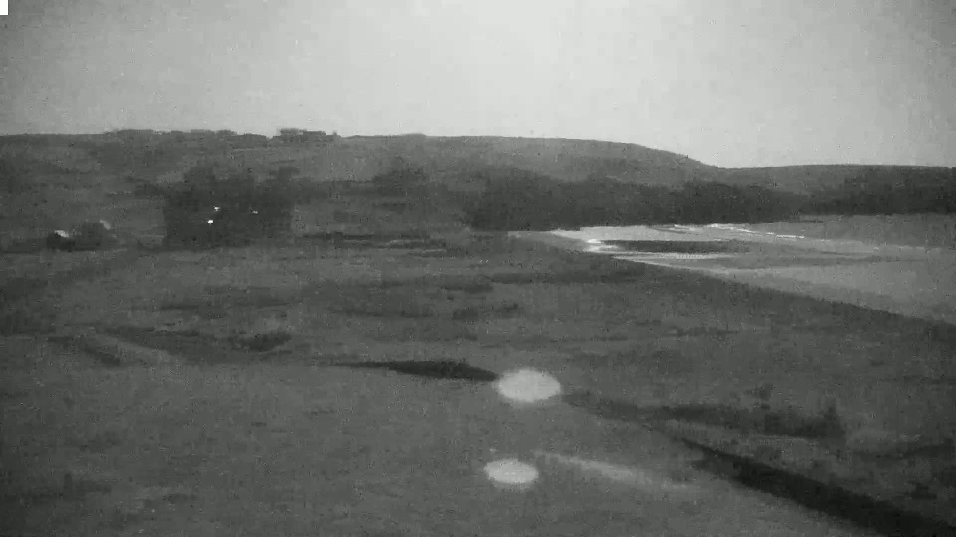 A dark, overcast sky looms over a flat, sandy shoreline near a low, treeline hill.