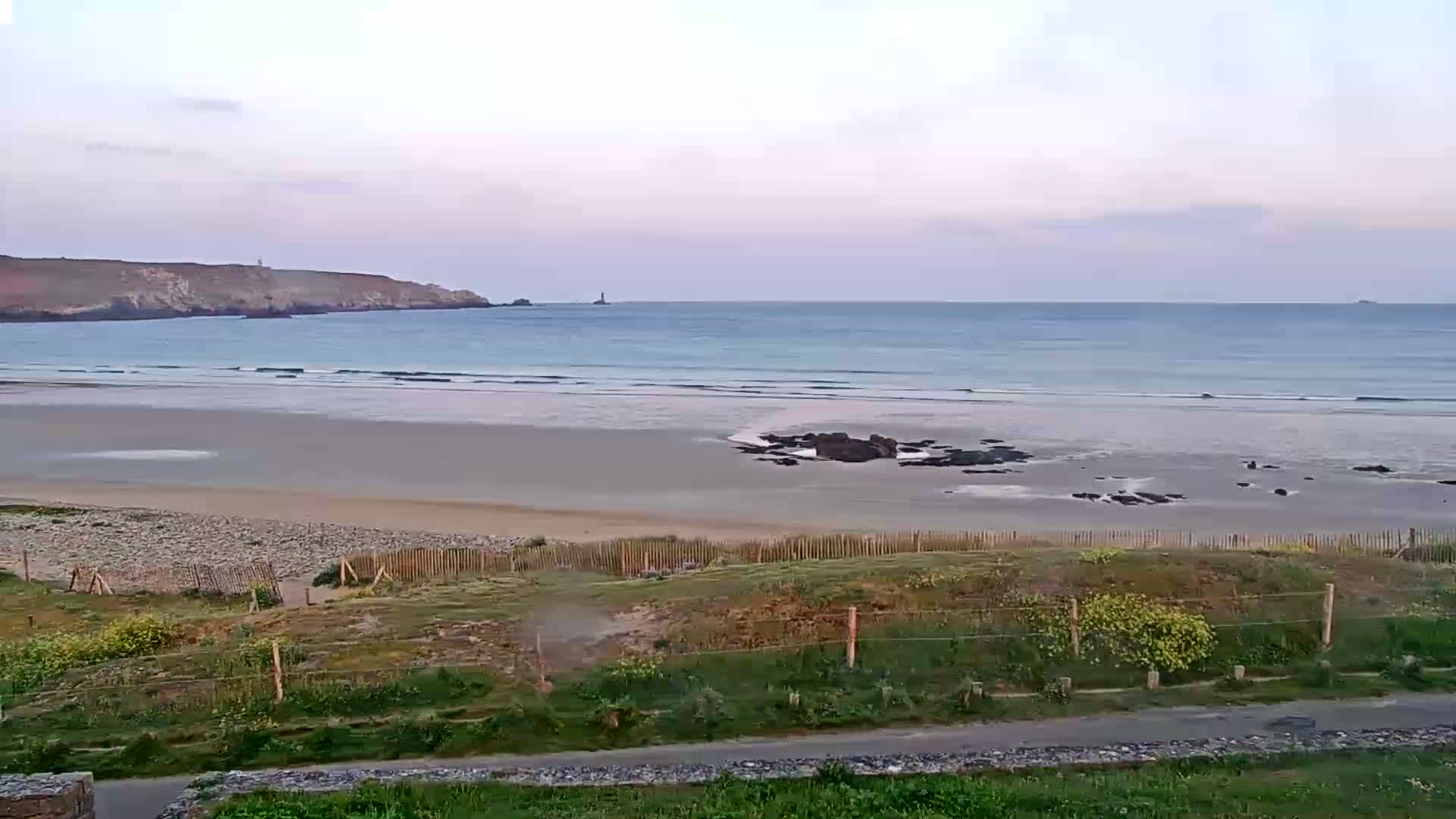 A sandy beach with gentle waves meets a calm ocean under a pale, overcast sky, backed by a low-lying coastline.
