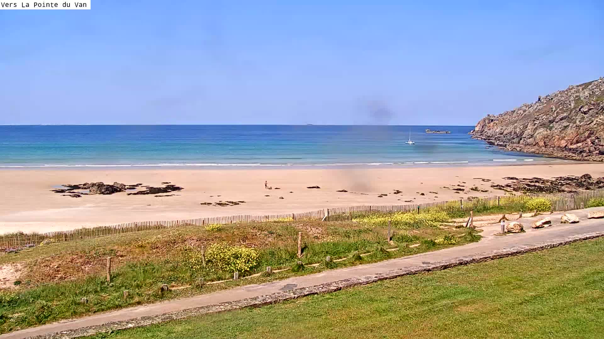 A sandy beach with calm, light blue water is partially enclosed by rocky cliffs under a clear, sunny sky.