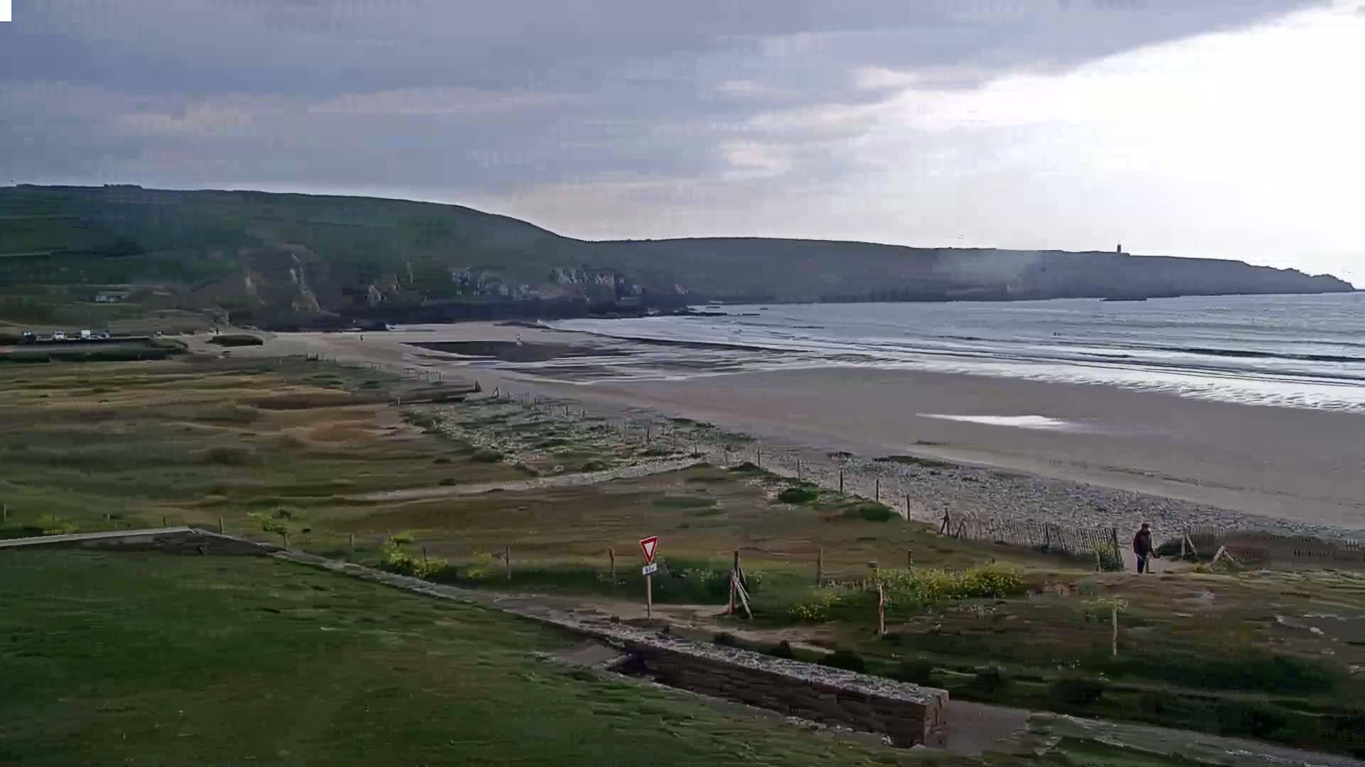 A sandy beach stretches along a coastline under a cloudy sky, with green hills in the background and a few people visible near the water's edge.
