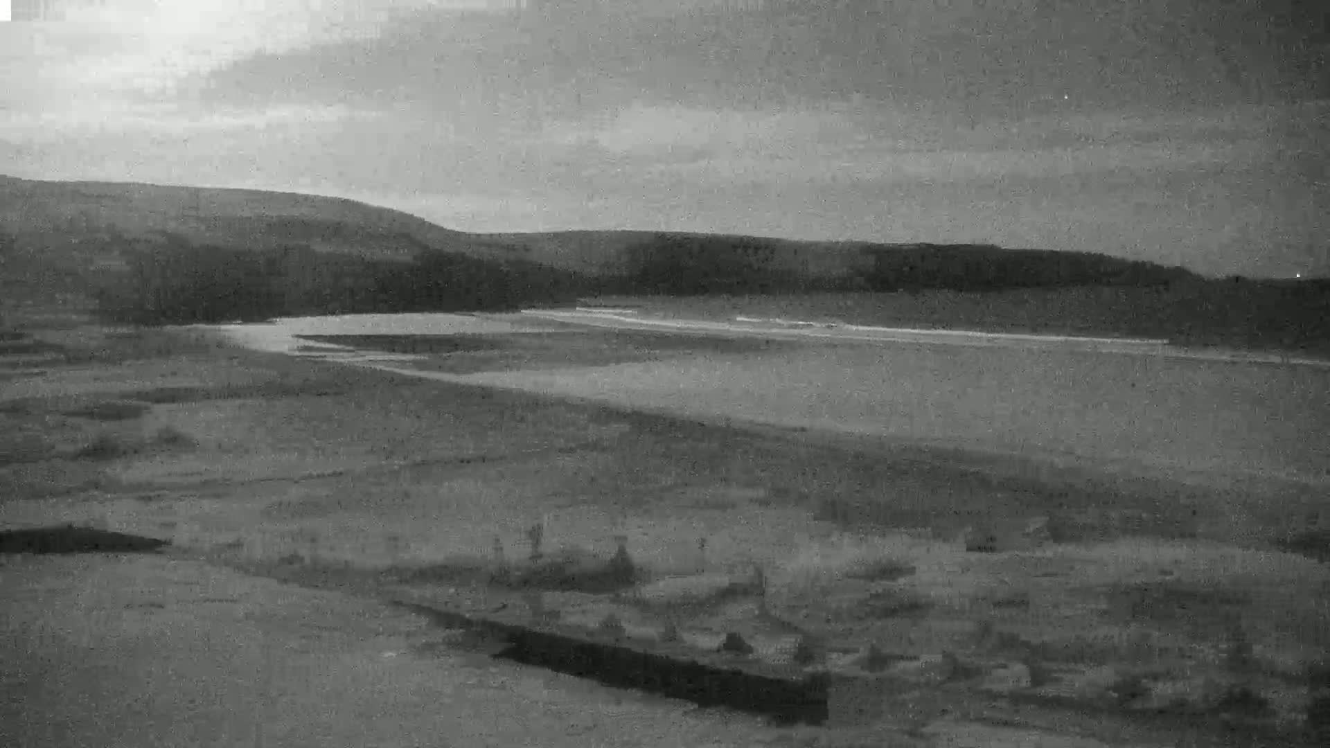 A grayscale image shows a calm body of water under a cloudy sky, with hills in the background and what appears to be a graveyard in the foreground.